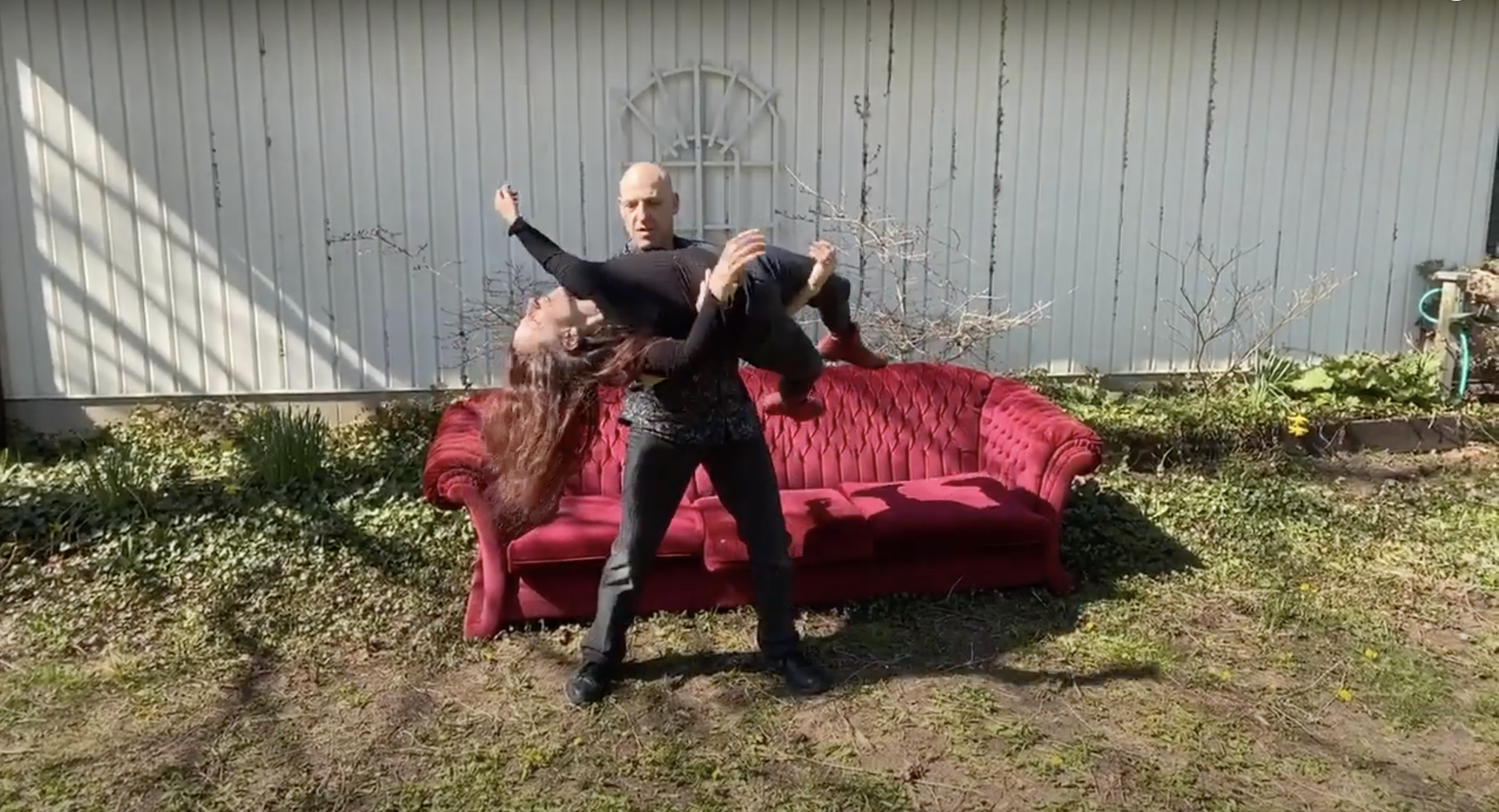 Two dancers, the male holding the female as she is laying in his arms, in front of a red couch outdoors