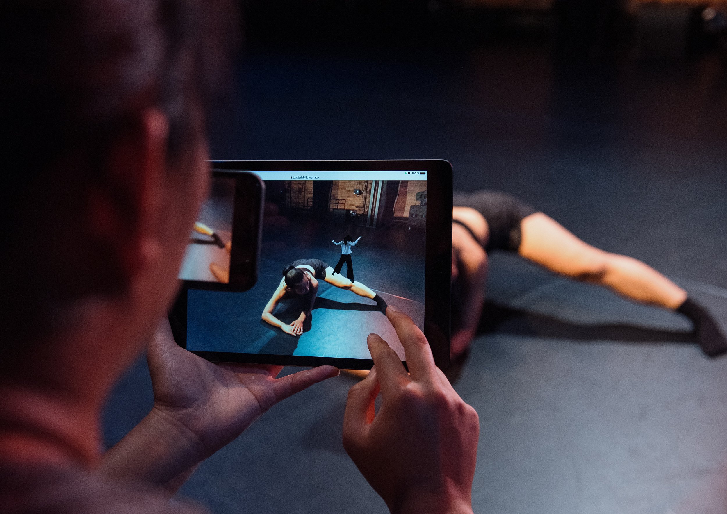  Mio Sakamoto, an East Asian woman in her mid 20s with long wavy hair, lunges to one side. A miniature version of Mio appears in Augmented Reality on an iPad screen standing on the real Mio’s leg. 