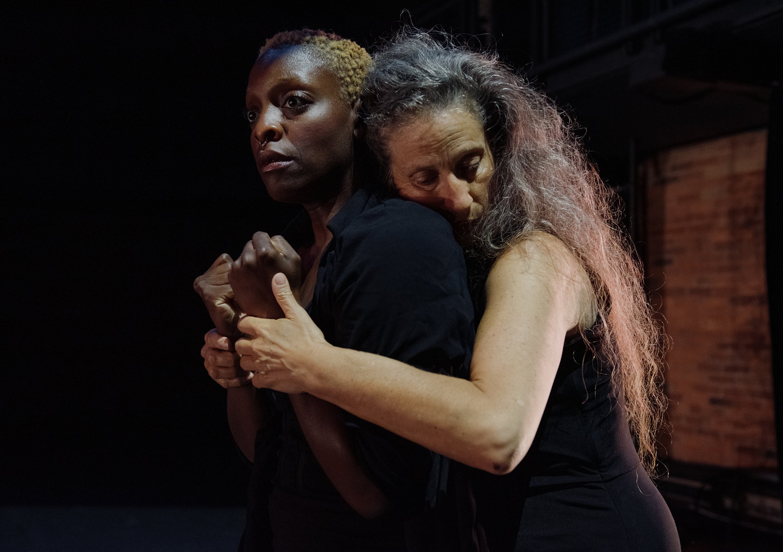  Karen Kaeja, an Ashkenazi white female with long grey hair gently embraces Nickeshia Garrick, a Queer, Afro-Caribbean Canadian artist with short kinky blonde hair. Karen has her eyes closed and holds Nickeshia’s upright fists. 