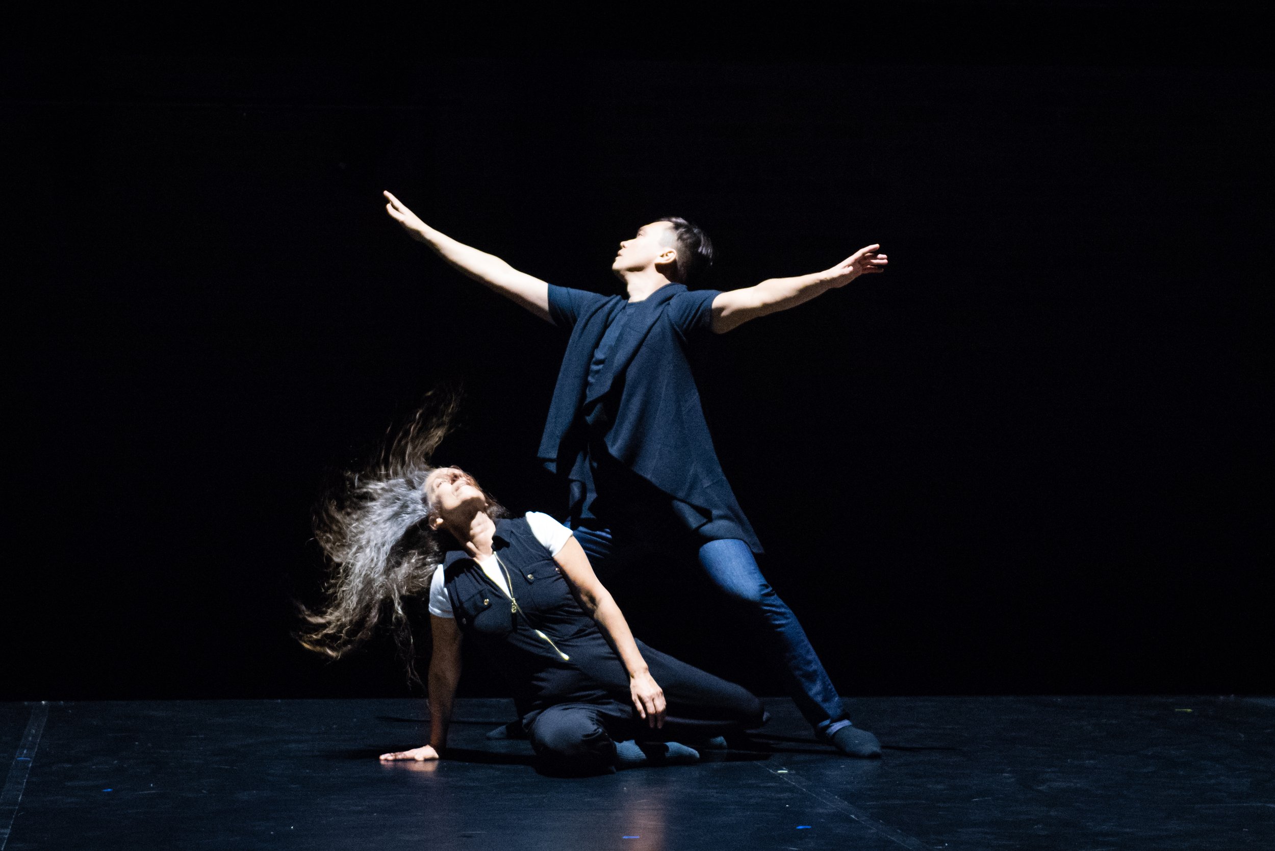  Karen Kaeja kneels on the floor with one hand bracing her and her hair flying out to one side. Michael Caldwell stands directly over her with his legs wide and his arms extended to either side. 