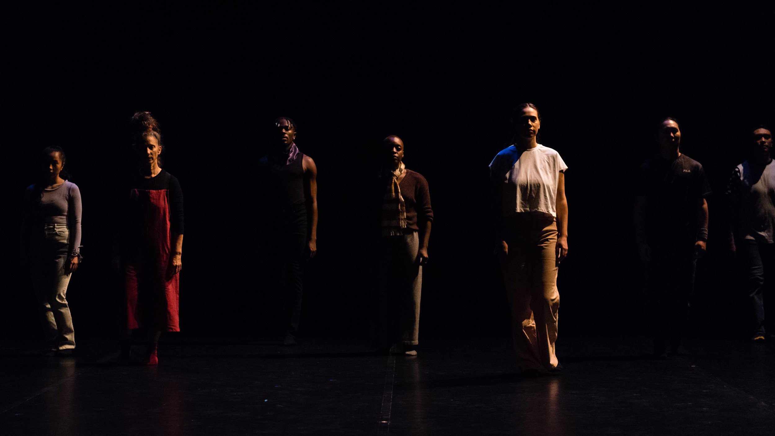  The full cast of I am the Child of… walk forward in a straight line that extends across the stage horizontally. The lighting is dark and shadowy. The performers have serious facial expressions and are diverse in age, gender, and ethnicity. 