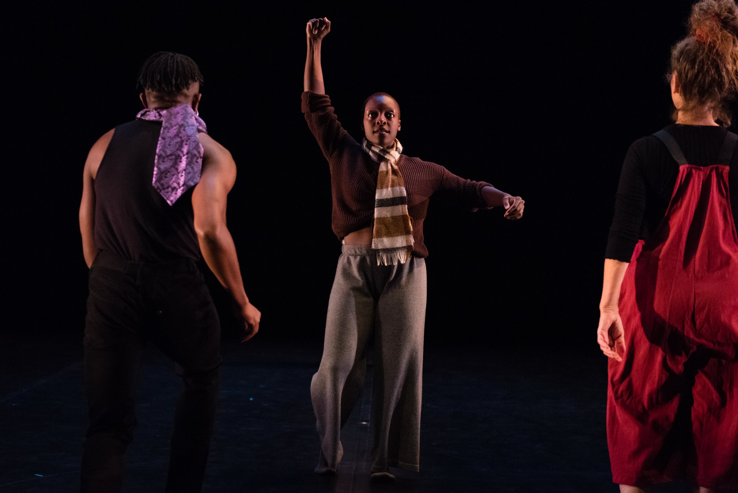  Nickeshia Garrick, a Queer, Afro-Caribbean Canadian artist with short kinky brown hair, extends one arm overhead and one to the side. Both hands are curled into fists. Two other performers approach Nickeshia with their backs to the camera. Nickeshia