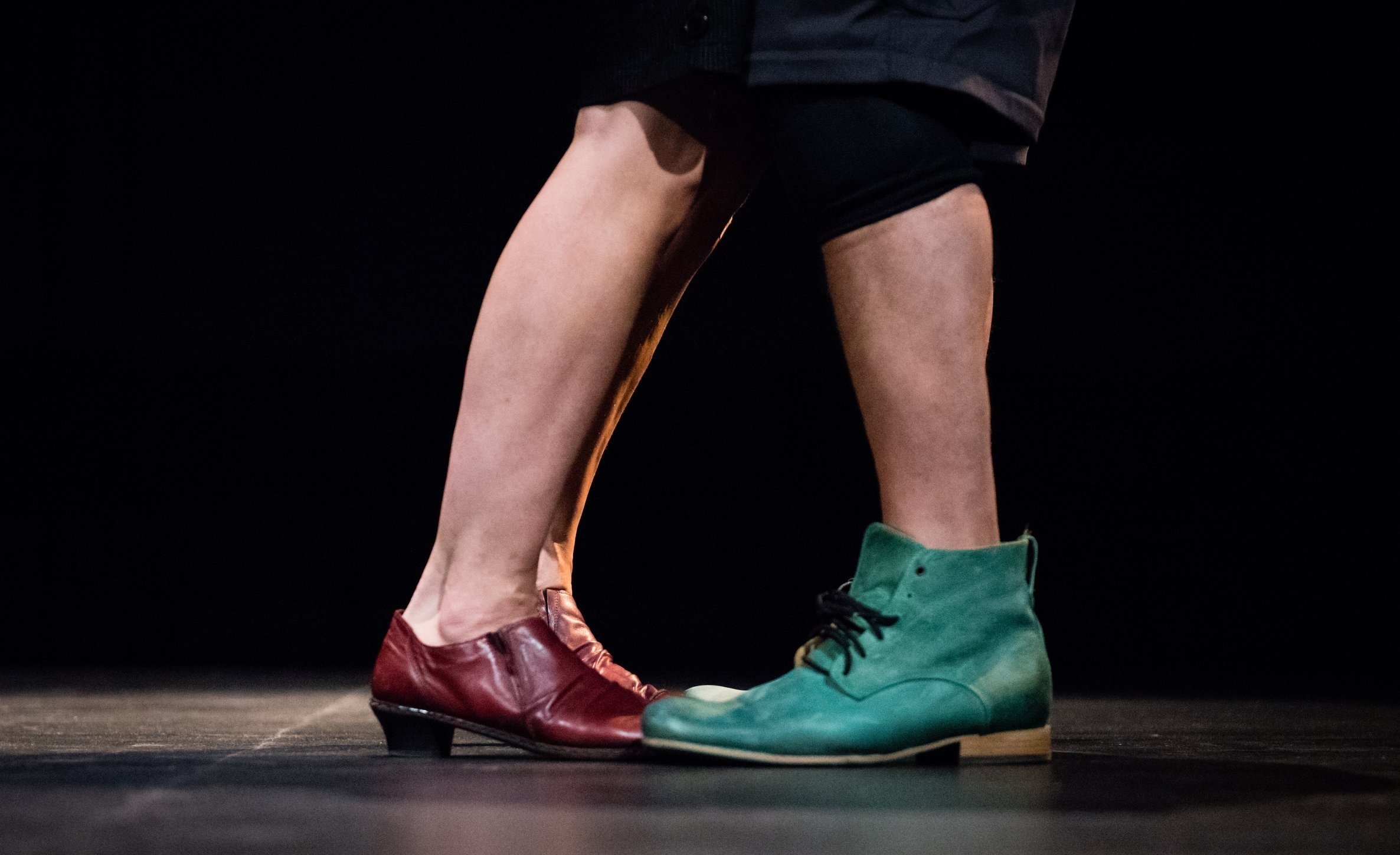 Two pairs of feet, one wearing red shoes and one wearing green shoes, stand toe to toe.