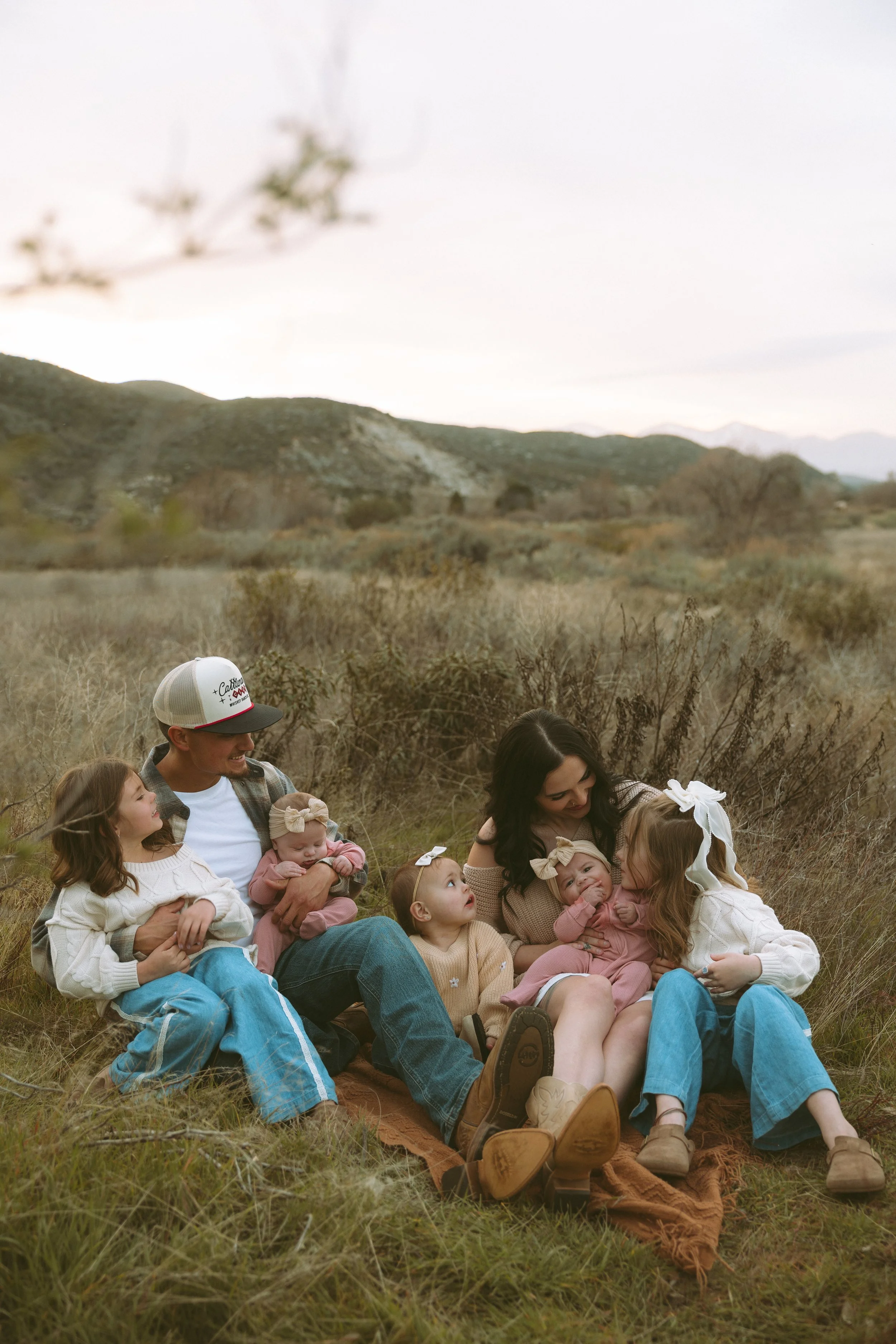 A family enjoying outdoor time in a park, with a man lifting a smiling baby girl in the air, a woman watching and smiling, and some children sitting nearby, surrounded by trees and sunny weather.