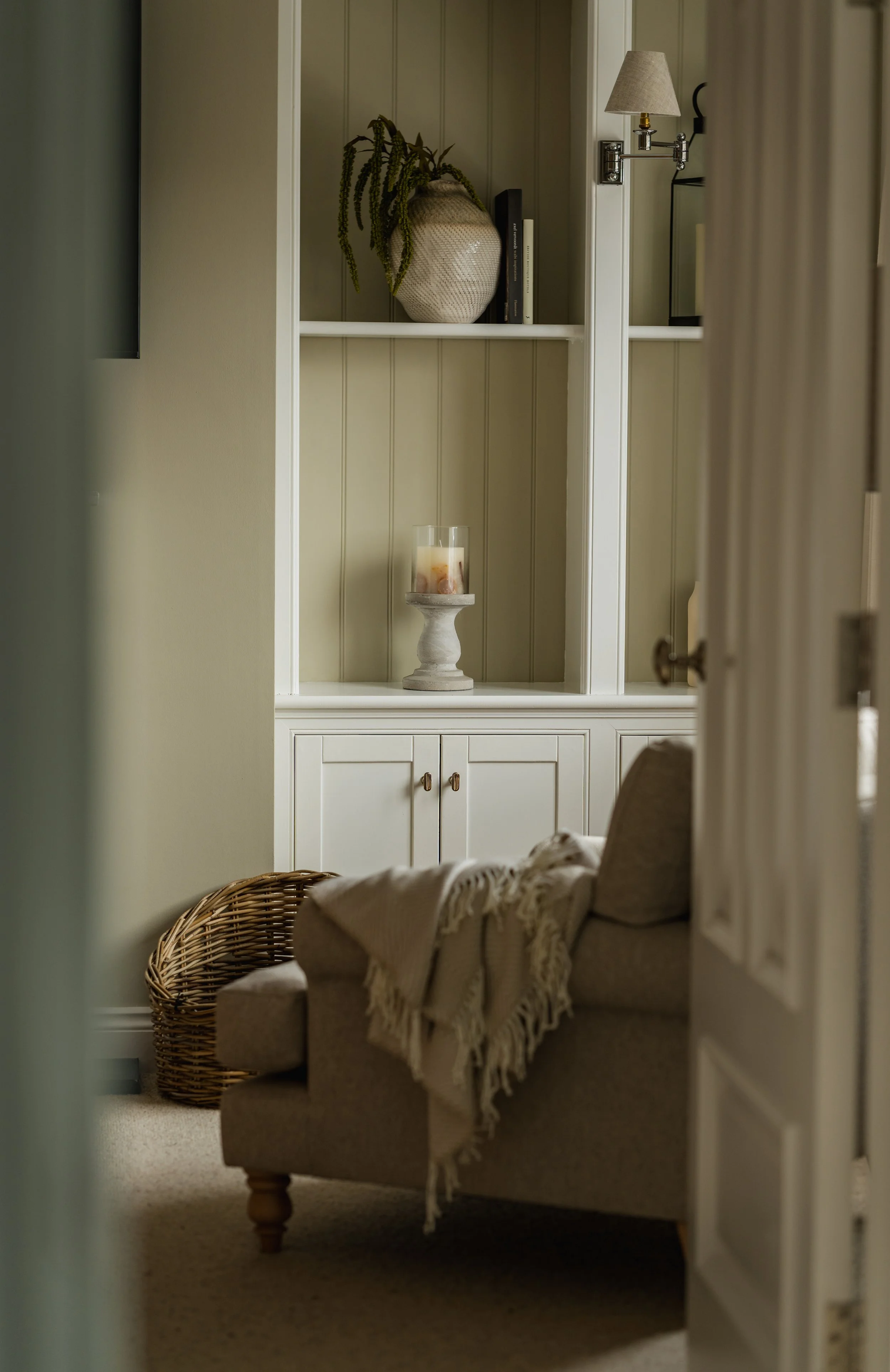 View of a cozy corner with a beige armchair covered by a white throw blanket, a woven basket to the side, and a white shelving unit with decorative items including a large vase with greenery, books, and a candle holder.