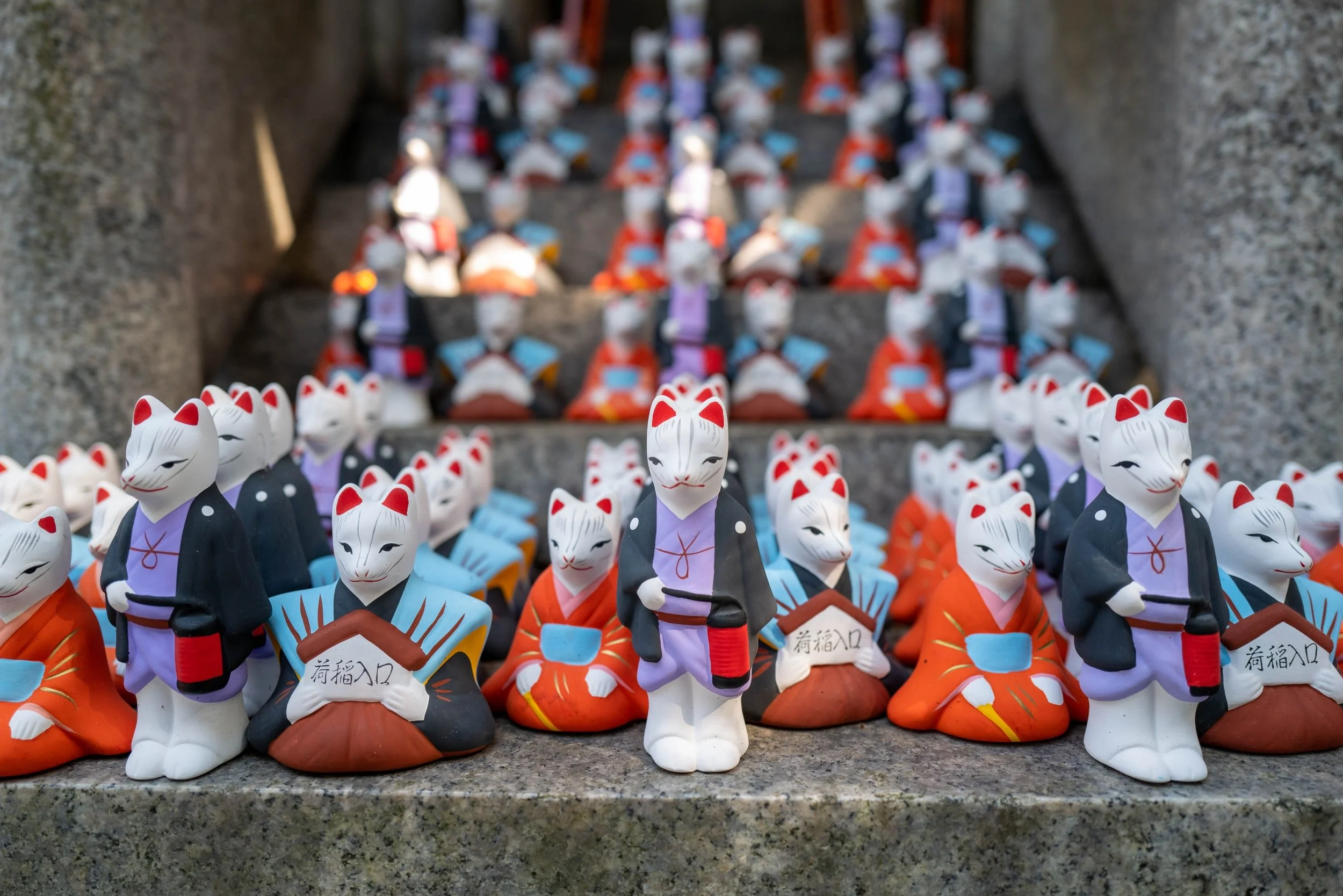 Anthropomorphic Maneki-neko wearing traditional clothing stand and sit in rows on stairs. The cats furthest away are blurry.
