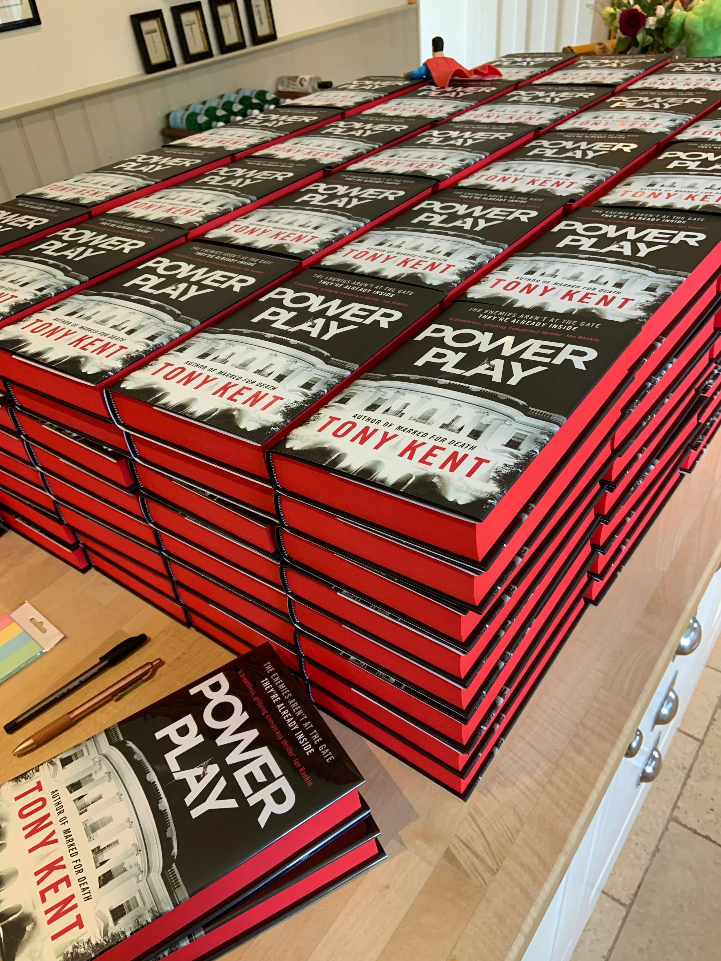 Stack of books titled 'Power Play' by Tony Kent on a table, with a few copies open and writing tools nearby.