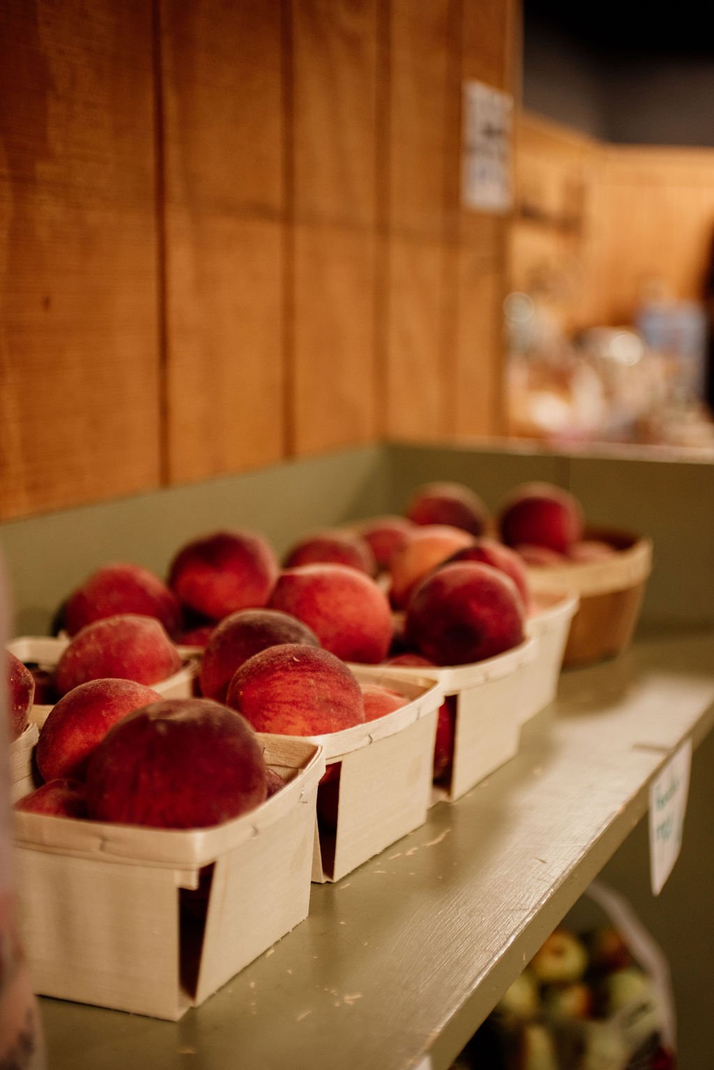 Fresh peaches in small wooden baskets on a metal shelf in a grocery store with wooden wall paneling in the background.
