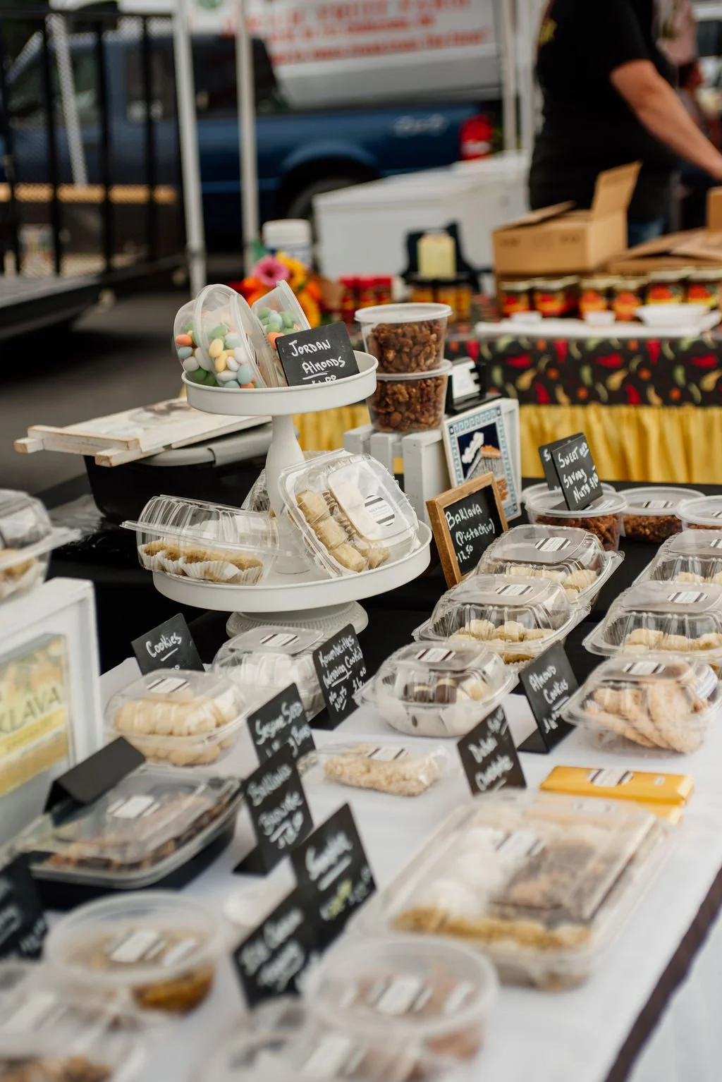 A market stall displaying various baked goods and snacks in plastic containers with black labels, including cookies, brownies, and baked treats, with a stand of Jordan almonds and packaged cookies on a three-tiered white display.