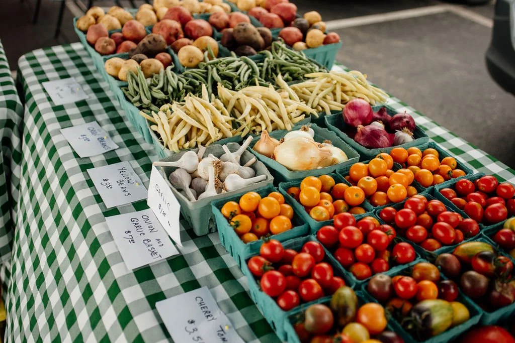 A farmer's market stall displays a variety of fresh vegetables, including tomatoes, garlic, onions, green beans, potatoes, and summer squash, arranged in teal baskets on a green and white checkered tablecloth.