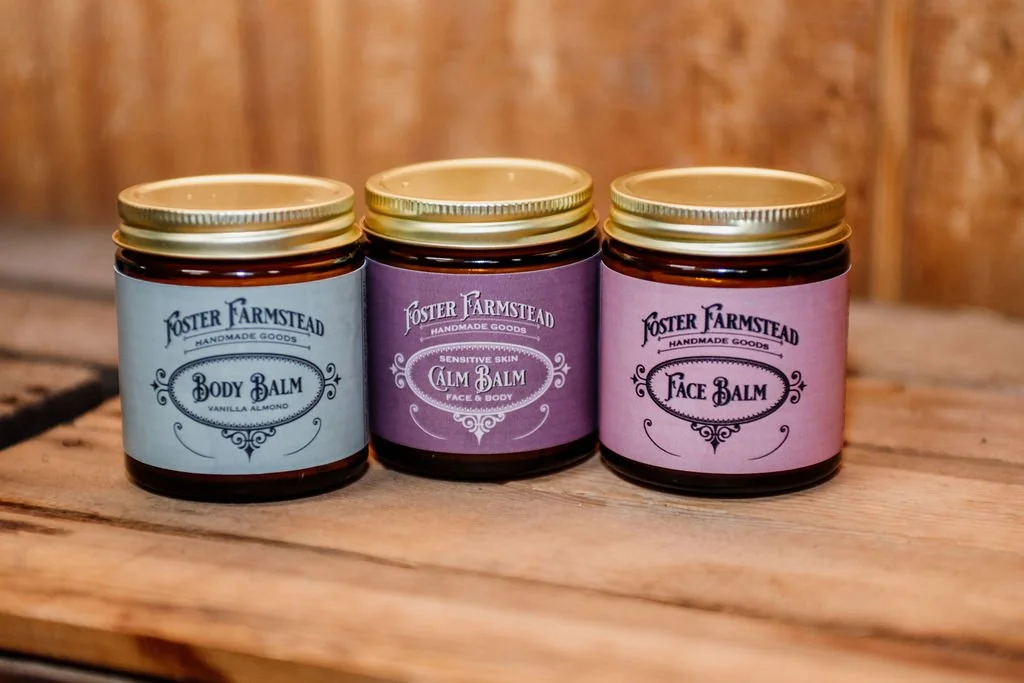 Three jars of Foster Farmstead handmade balm on a wooden surface, labeled 'Body Balm', 'Calm Balm', and 'Face Balm' in blue, purple, and pink labels, respectively.