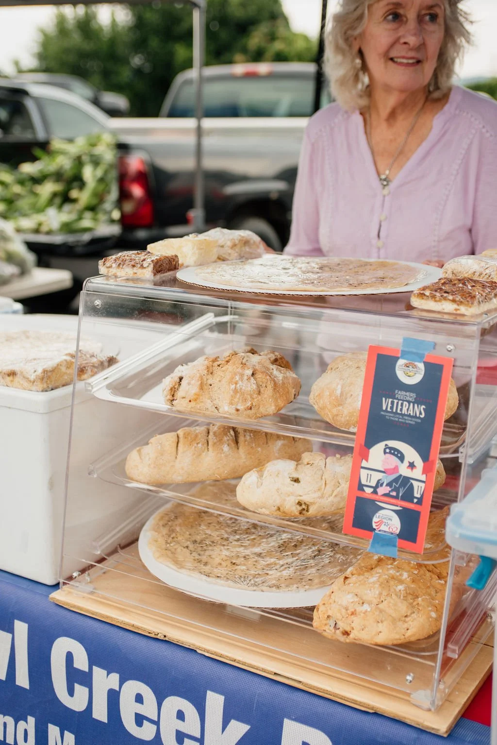 A woman smiles behind a display of baked goods including bread, a pie, and cookies at a market stall. There are cars and greenery in the background.