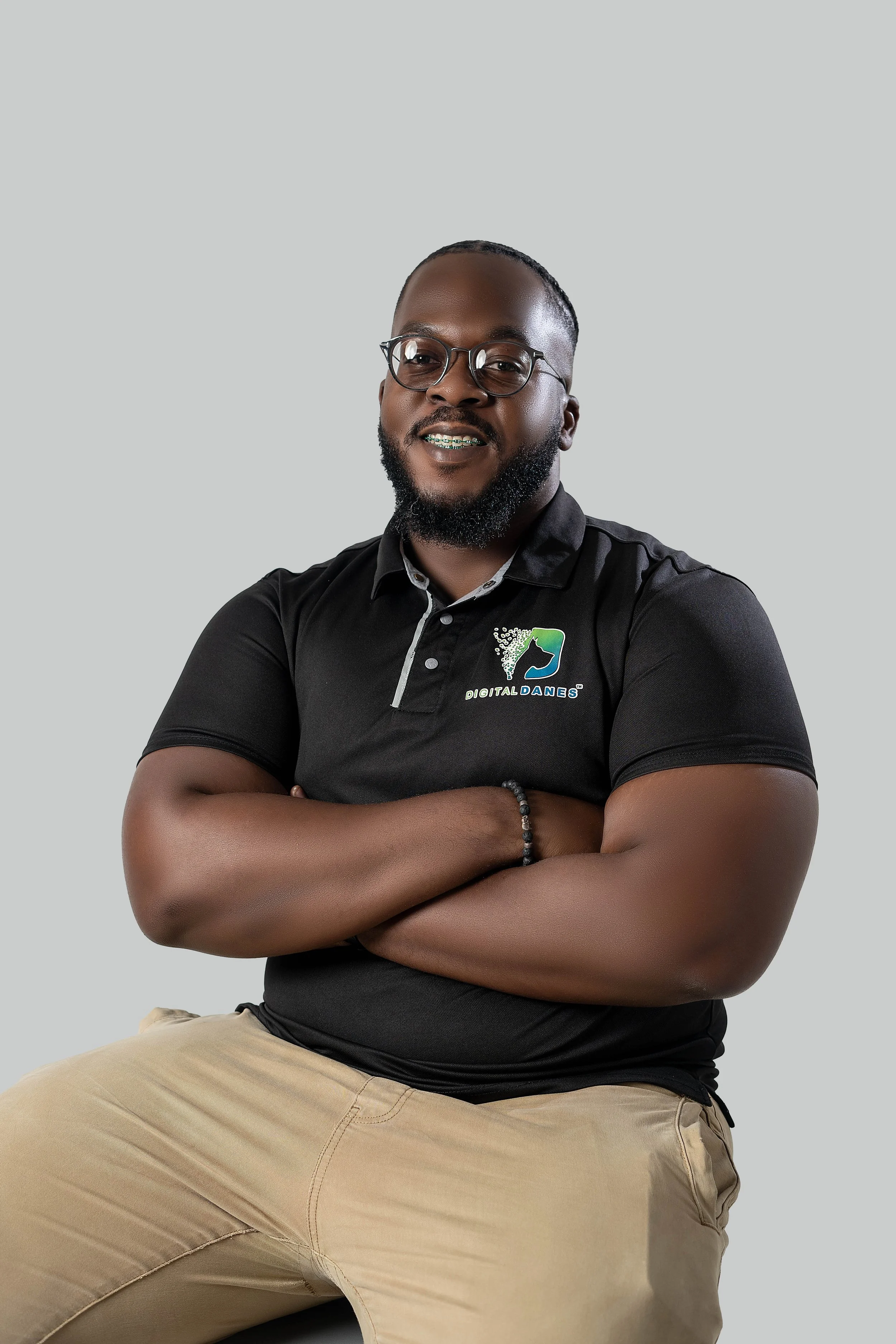 A man with glasses and a beard, wearing a black polo shirt with a logo and text 'DIGITAL DANES,' is seated with arms crossed against a plain light gray background.
