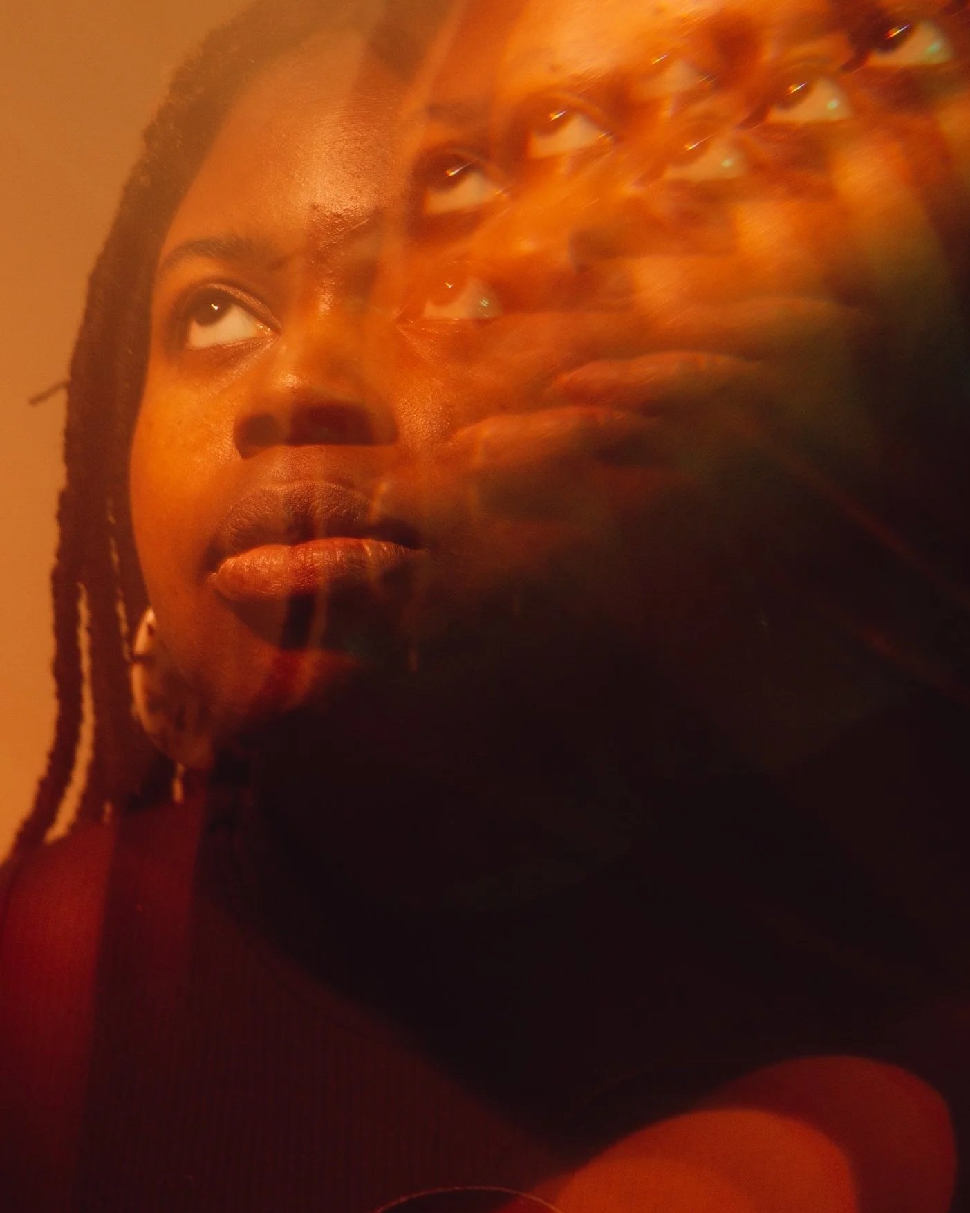 Image of Diana. They are a Black person with thick braids looking up and to the left. There is an effect on the camera lens that creates overlapping images of their face
