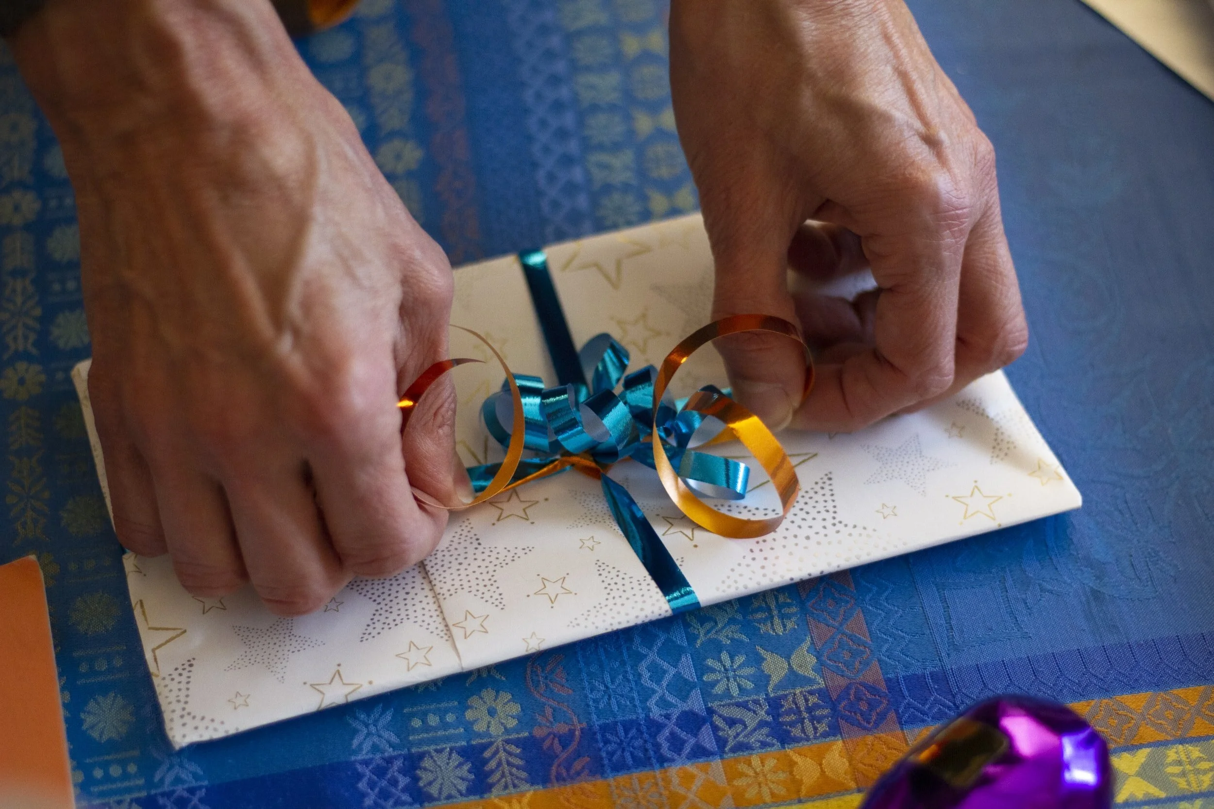 Hands wrapping a gift with white paper decorated with gold star patterns and blue and orange curled ribbons.