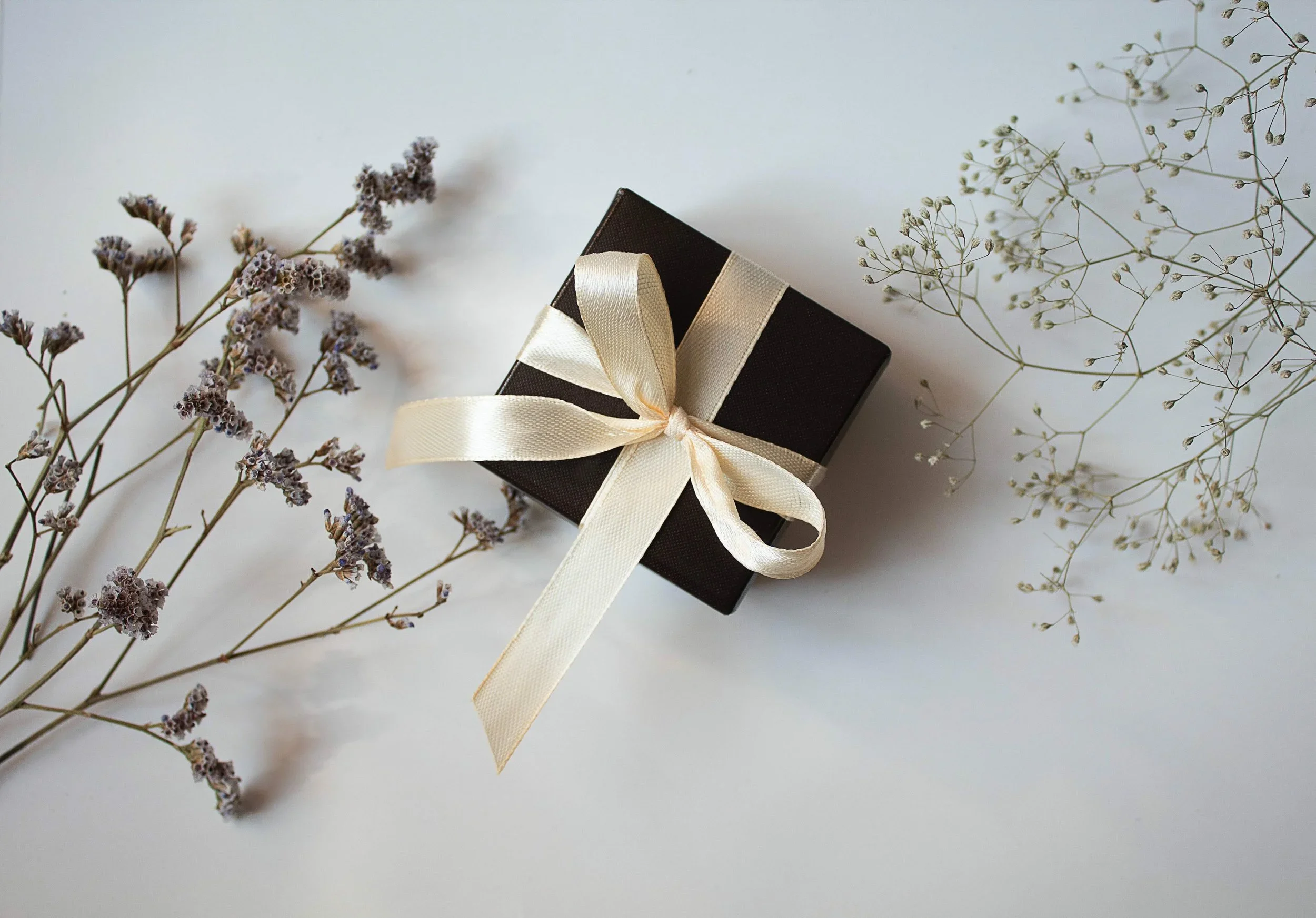 A small black gift box wrapped with a cream-colored satin ribbon tied in a bow, placed on a white surface with sprigs of purple and white dried flowers around it.