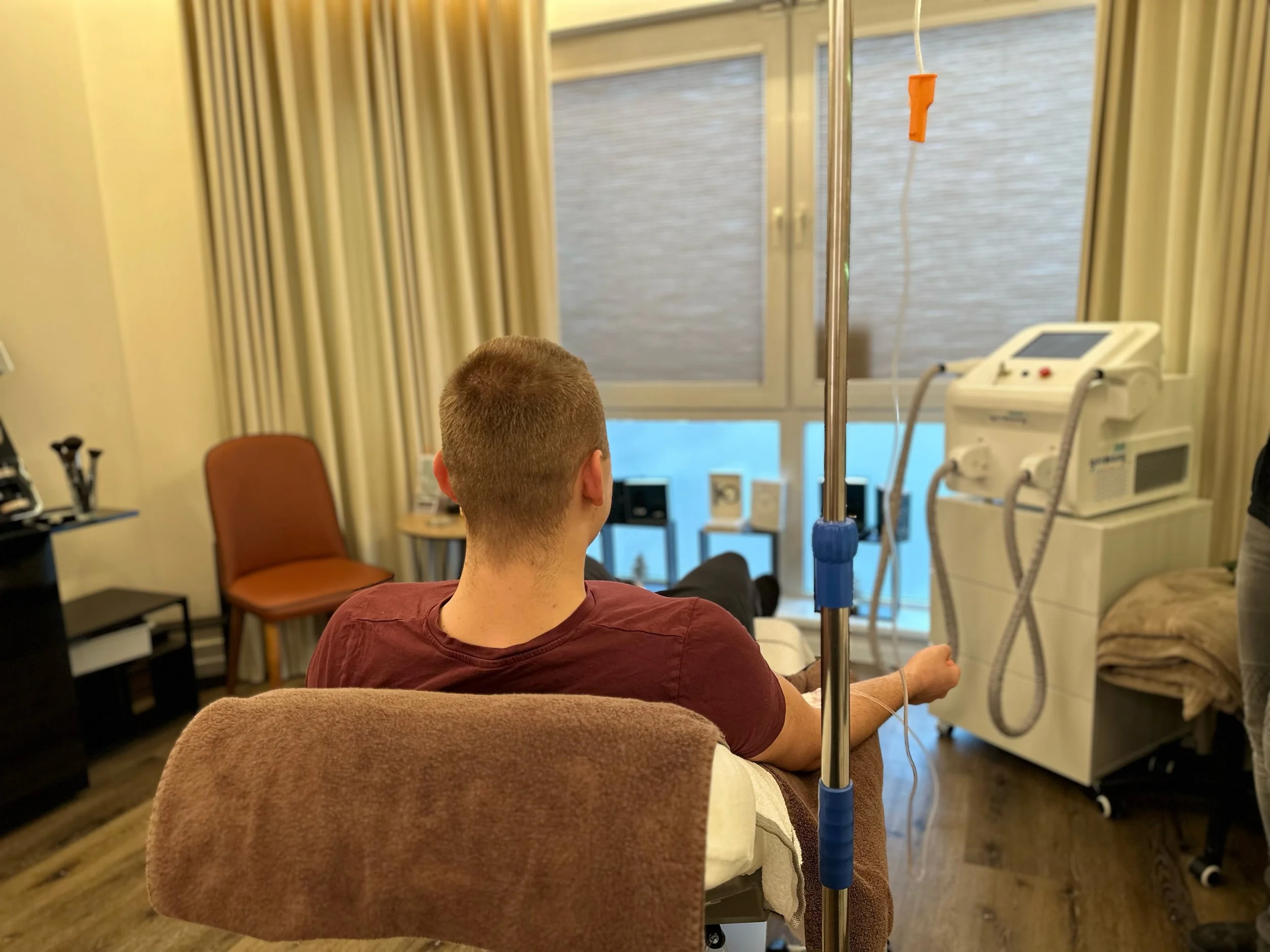 Ein Mann sitzt in einem medizinischen Behandlungsraum an einem Tropf. Er trägt ein dunkelrotes T-Shirt und sitzt auf einem Stuhl mit einem Handtuch über der Rückenlehne. Im Hintergrund sind ein Fenster mit Vorhängen, ein leeres Stuhl und ein medizinisches Gerät zu sehen.
