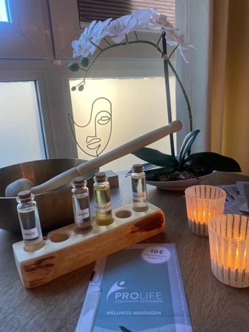 Innenaufnahme eines Entspannungsraums mit Aromatherapie-Elementen. Zu sehen sind eine Klangschale mit einem Klöppel, vier Fläschchen mit ätherischen Ölen in einem Holzständer, zwei brennende Kerzen und eine Broschüre. Im Hintergrund steht eine Orchidee auf dem Fensterbrett.