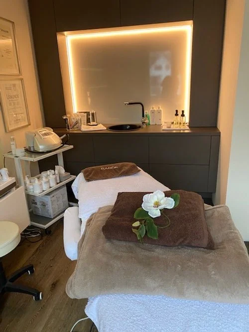 Massage- oder Behandlungszimmer mit einem Massagetisch, der mit Handtüchern und einer Blume dekoriert ist, sowie einem Waschbecken, Pflegeprodukte und Kosmetikgeräte im Hintergrund.