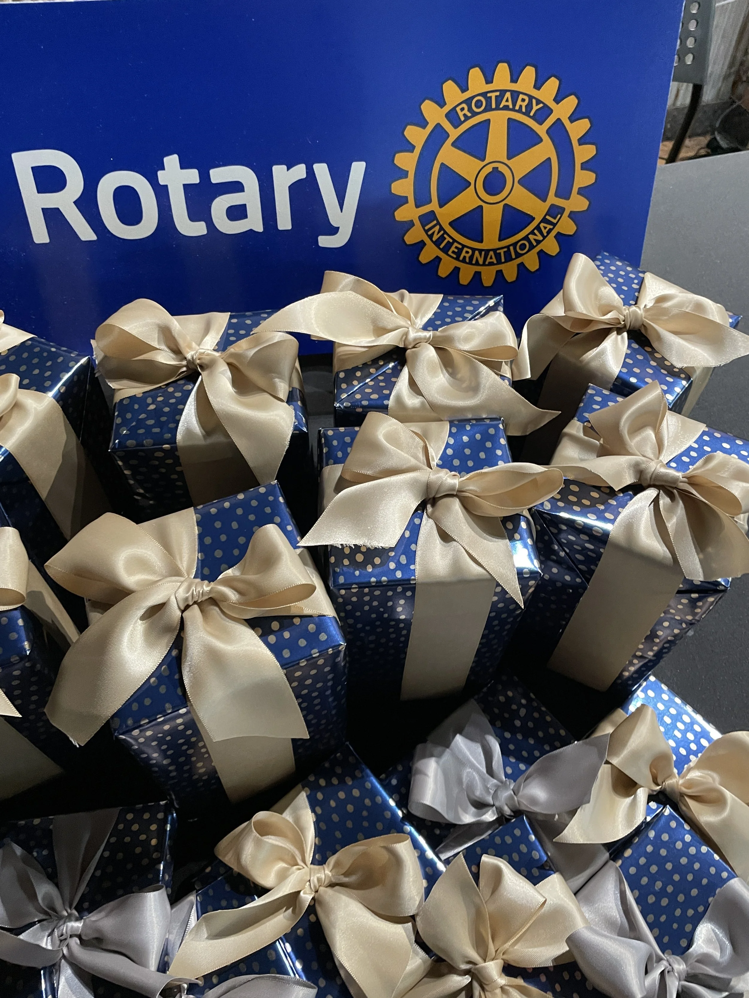 Multiple gift boxes wrapped in blue paper with white pearl dots, tied with cream-colored satin ribbons, placed in front of a Rotary International sign.