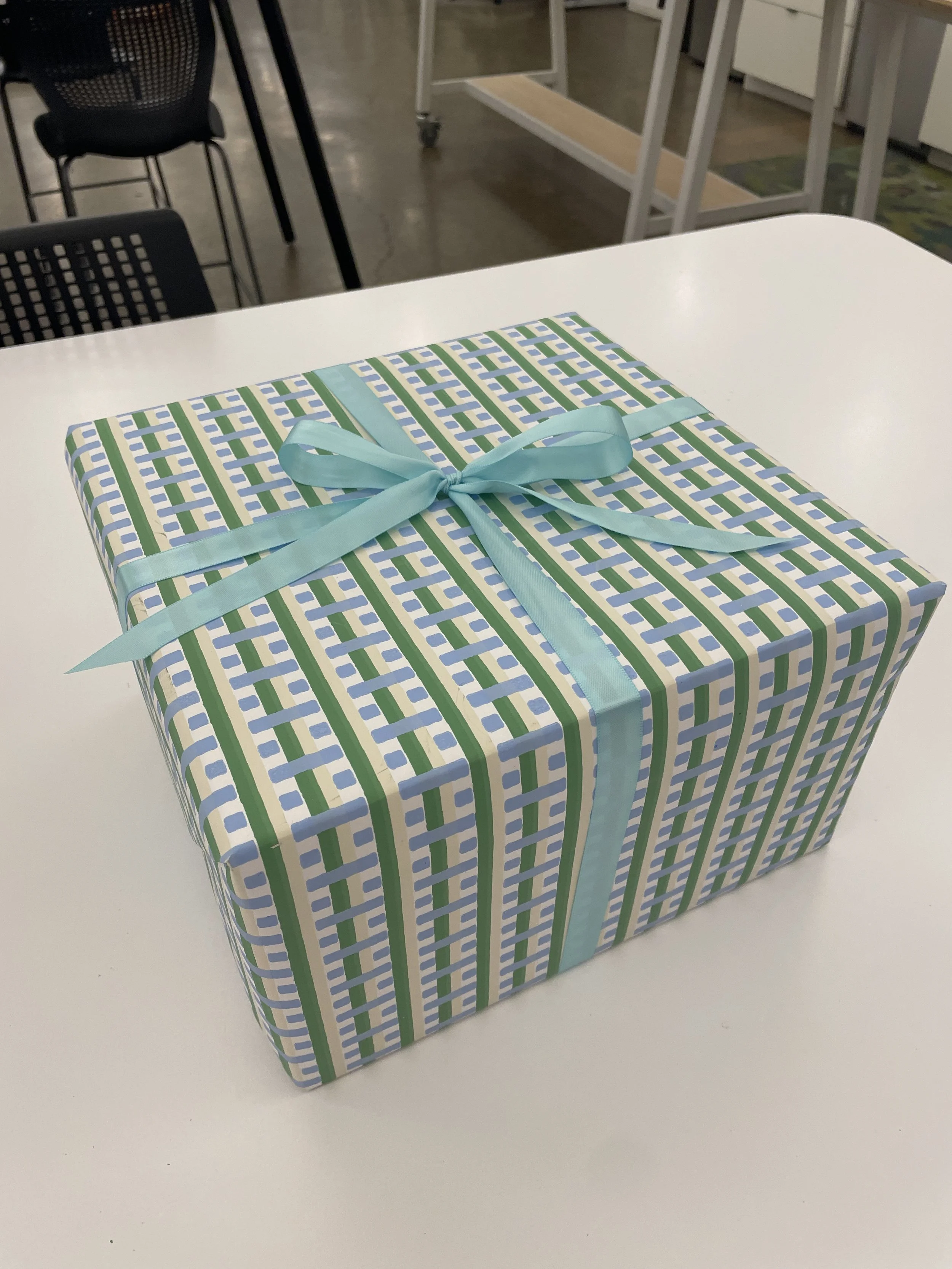 A gift box wrapped in blue, green, and white geometric patterned paper with a blue satin ribbon tied in a bow, placed on a white table.