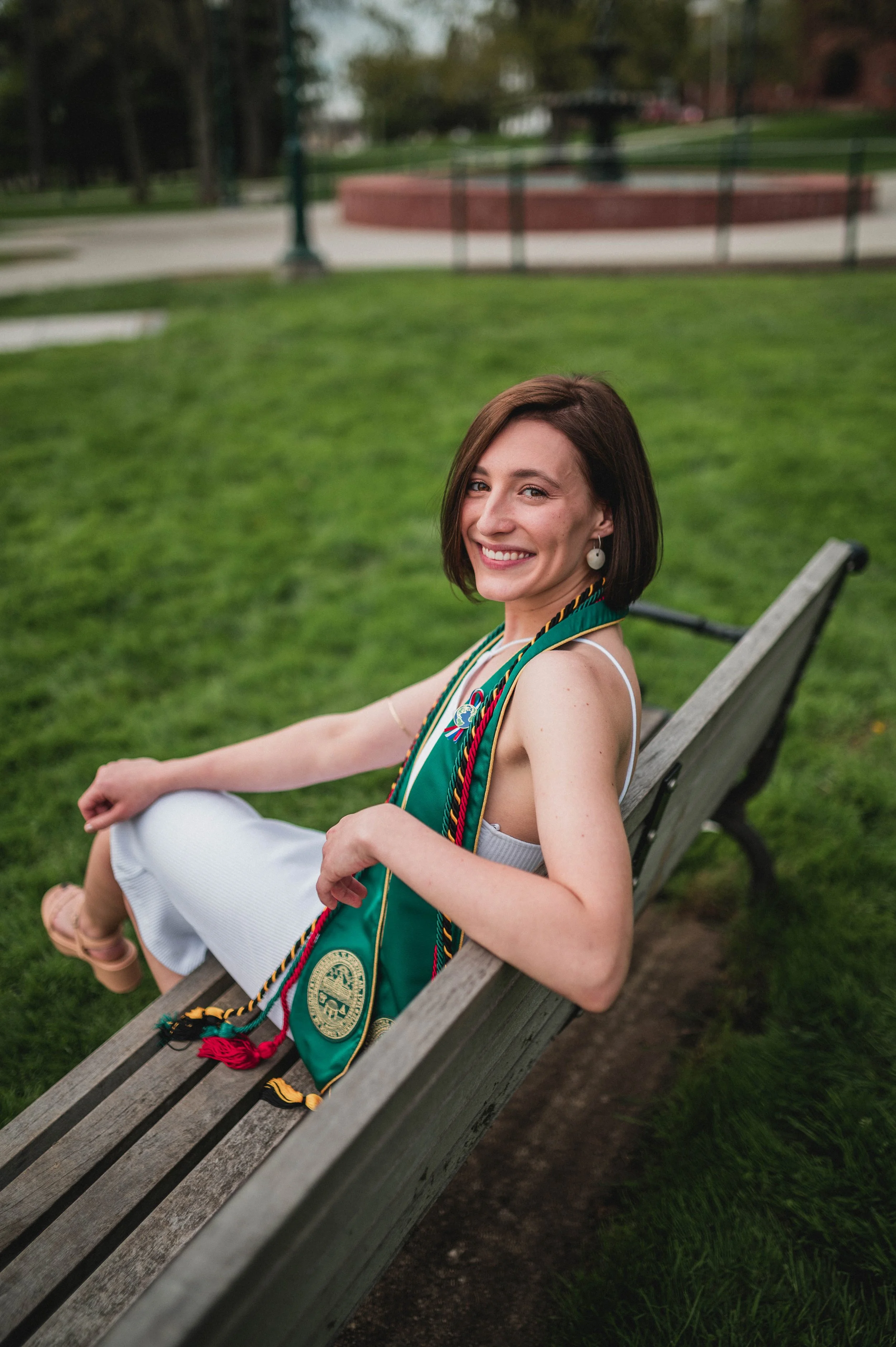 Woman seated on a bench outdoors, wearing graduation regalia with green and gold accents, smiling at the camera. Background shows grassy field and blurred fountain.