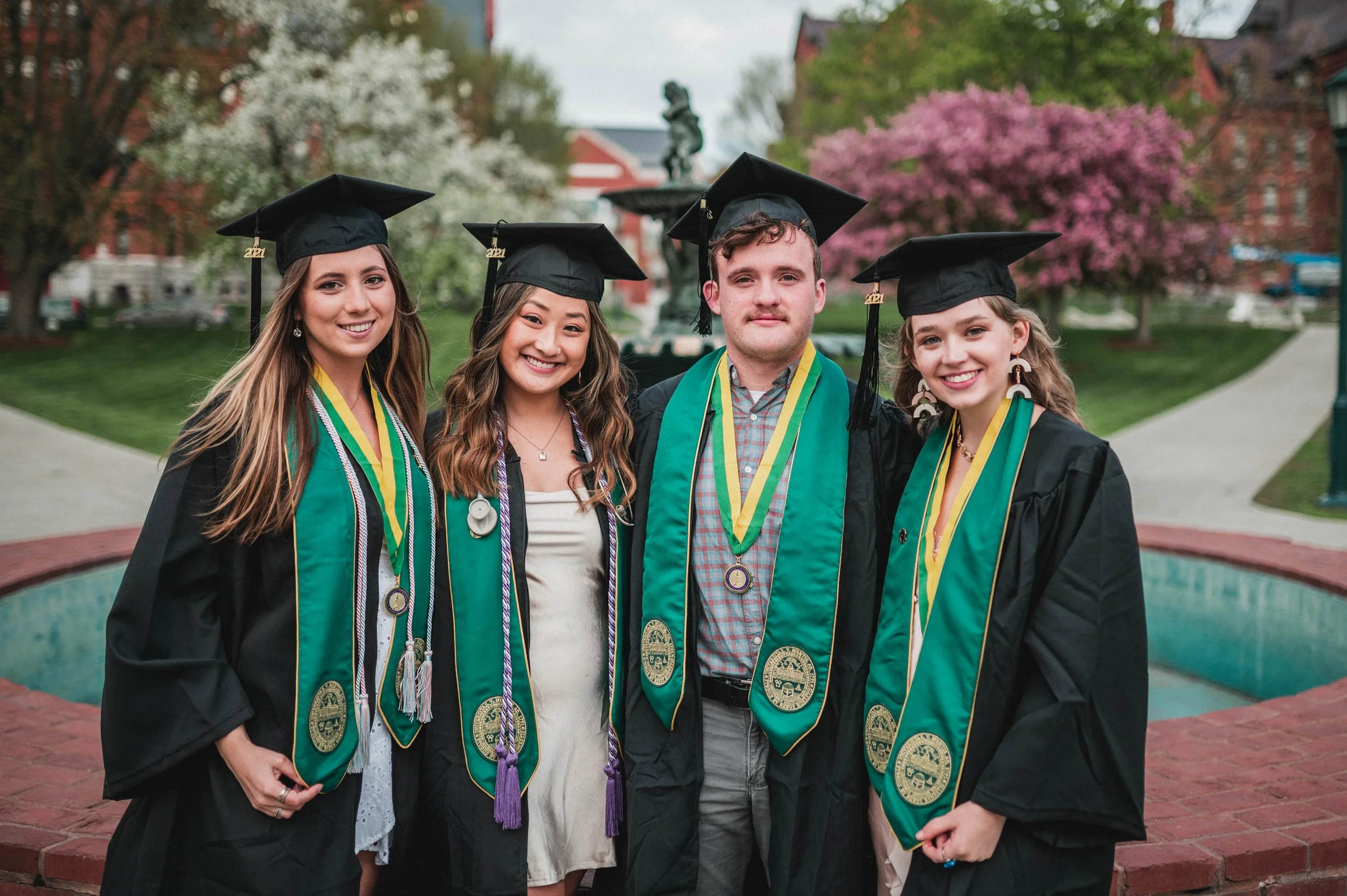 Four graduates in caps and gowns, smiling outdoors with blooming trees.