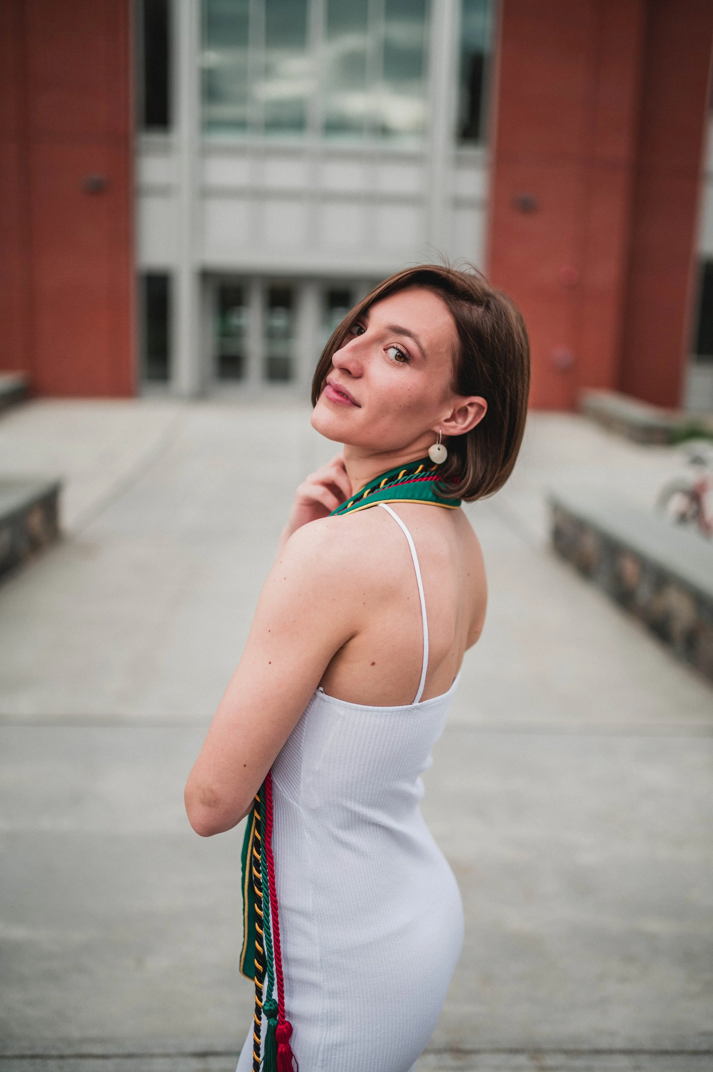 Woman in a white dress with green and red honor cords, standing outdoors, turning back to look at the camera.