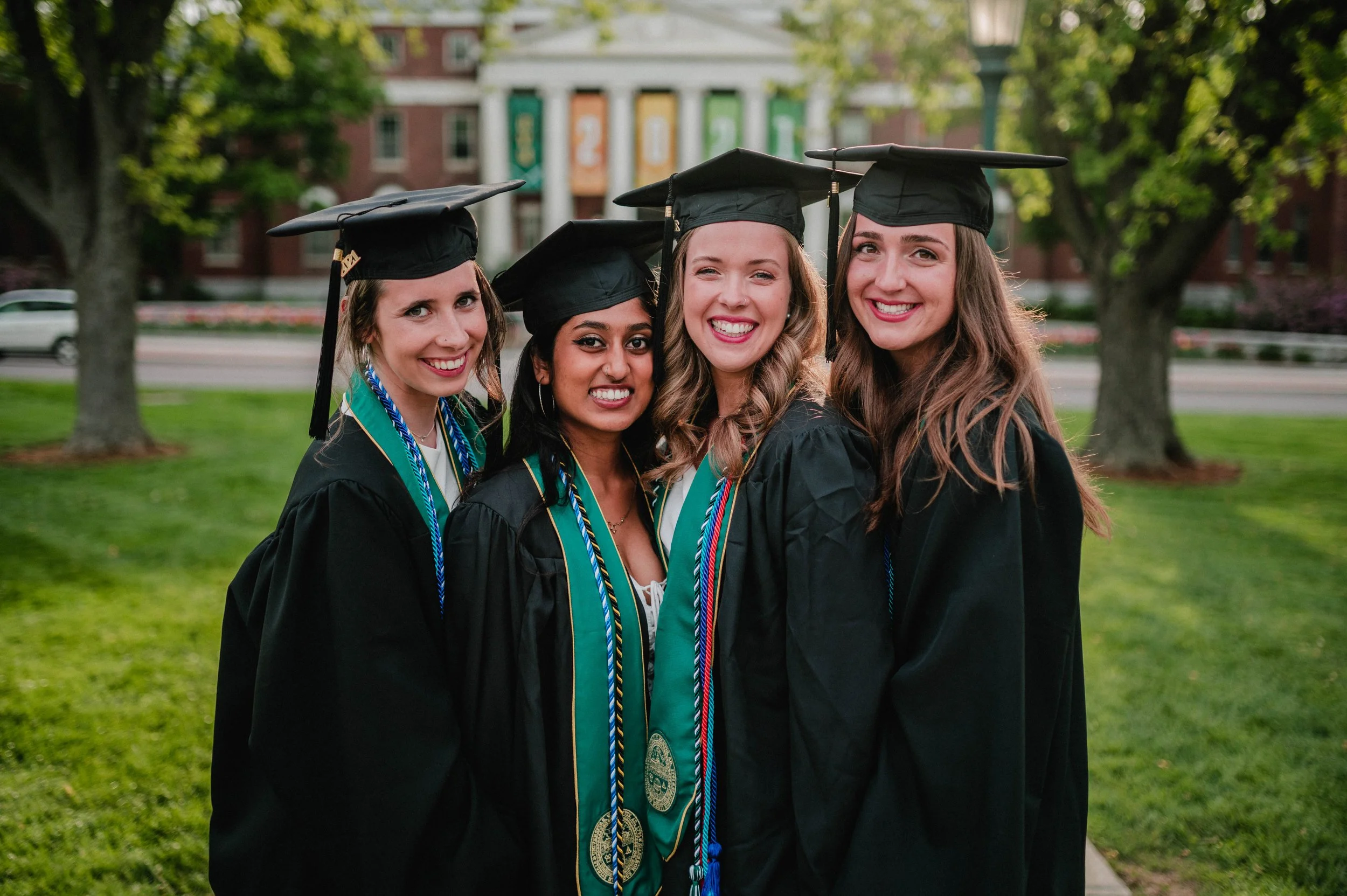 Four female graduates in caps and gowns smiling on a college campus.