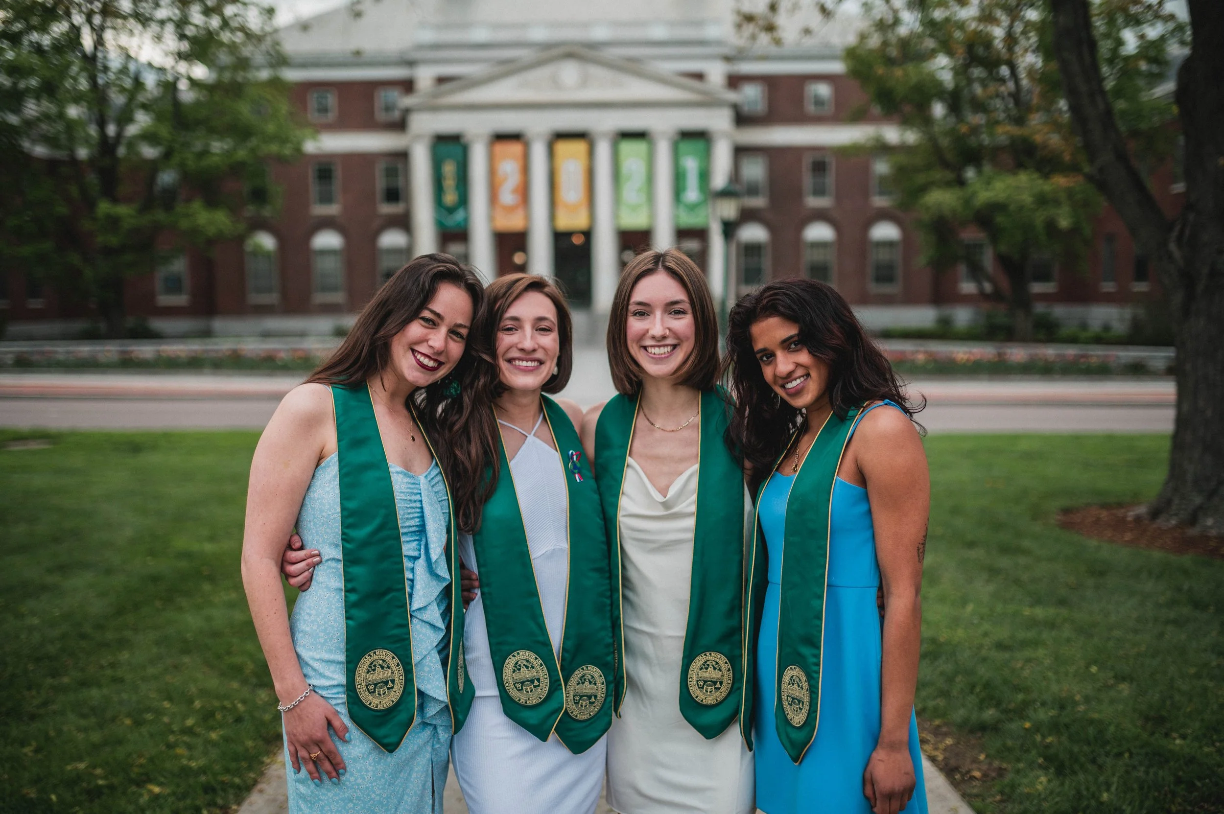 Four young women wearing graduation stoles stand together smiling in front of a building with 2021 banners.