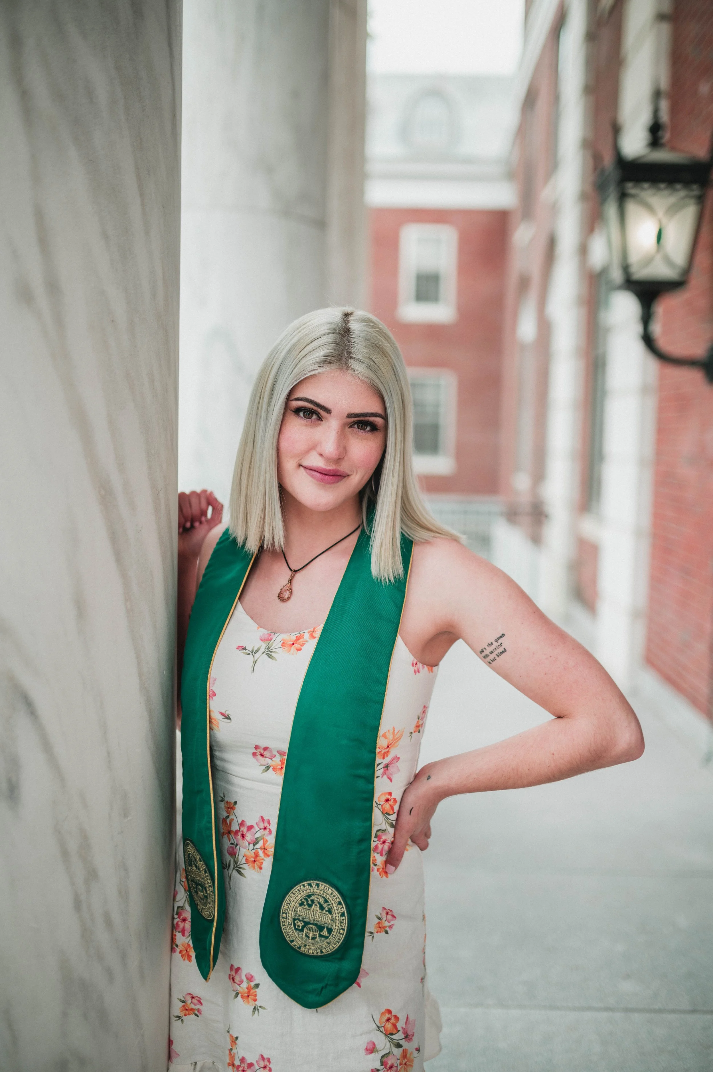 A young woman with blonde hair wearing a floral dress and a green graduation stole standing near a marble column on a university campus.