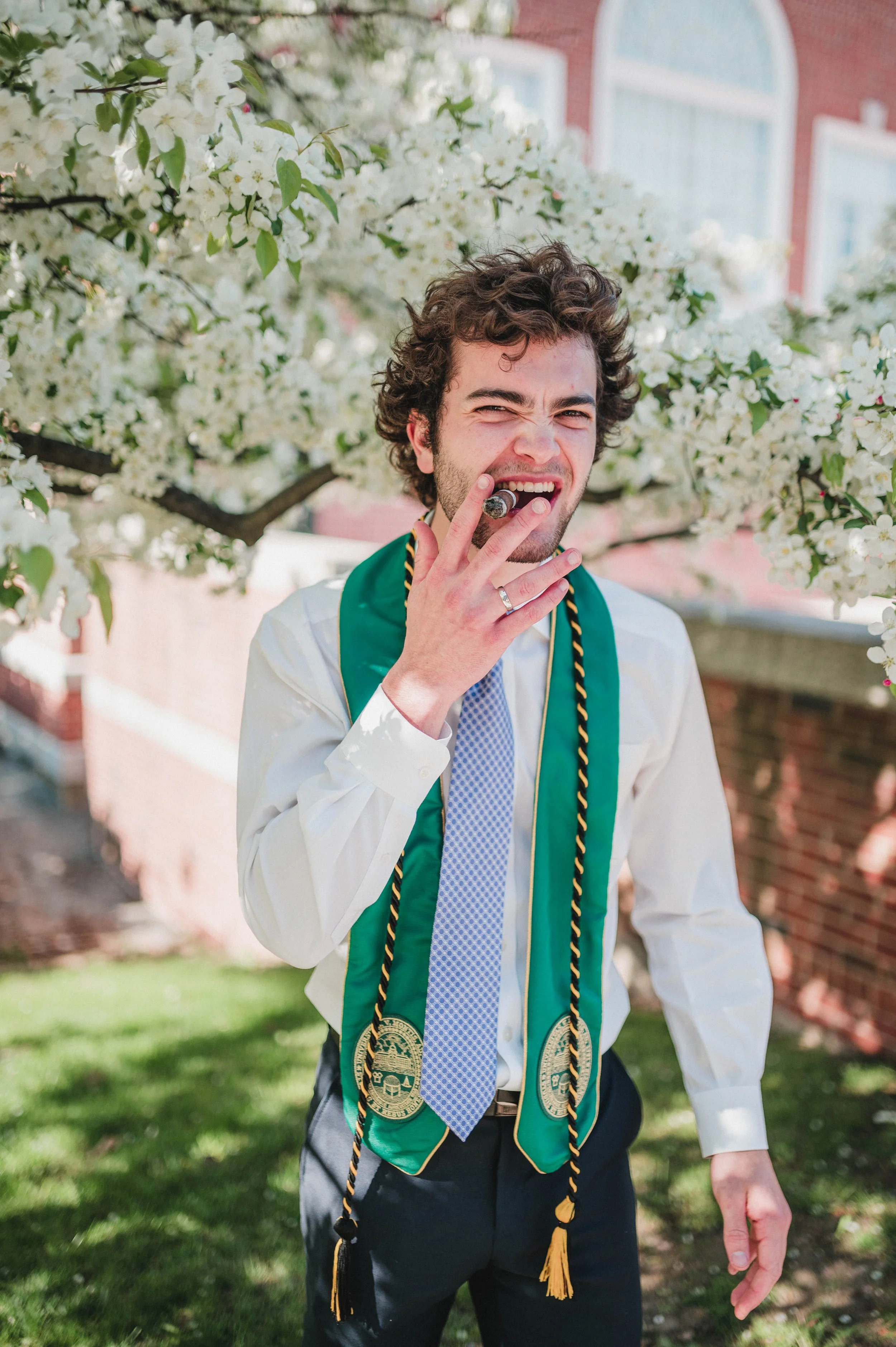 Man in graduation attire smoking cigar under blooming tree
