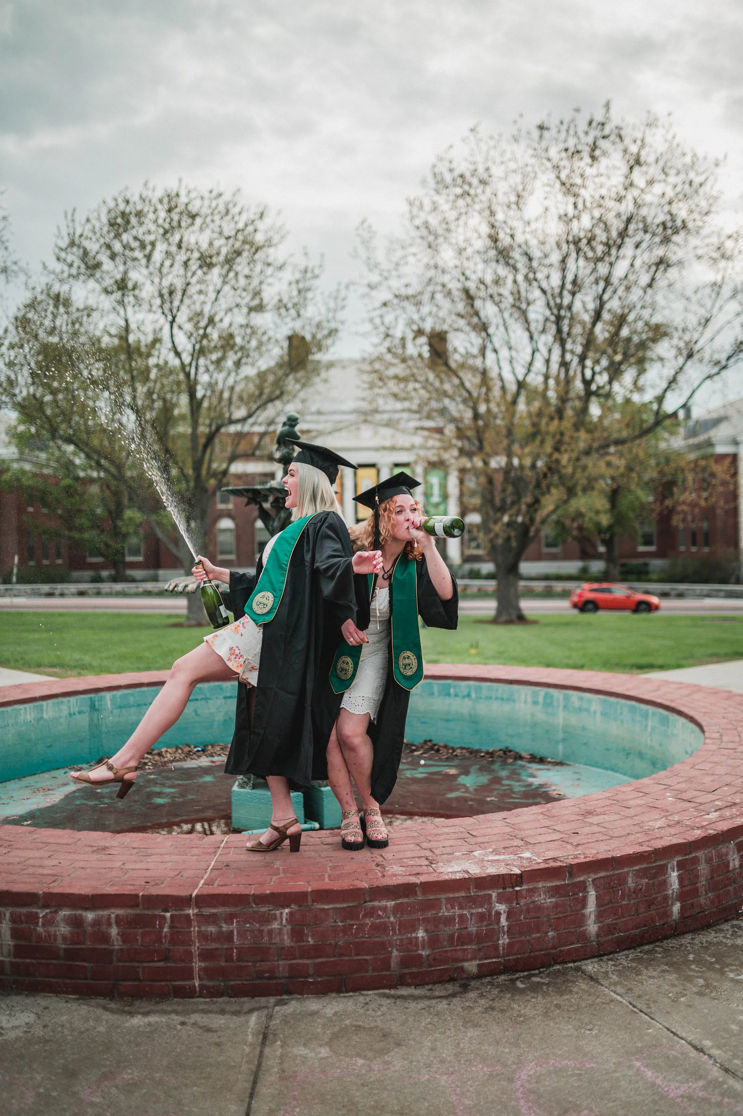 Two graduates in caps and gowns celebrate by a fountain, one spraying champagne and the other drinking from a bottle.