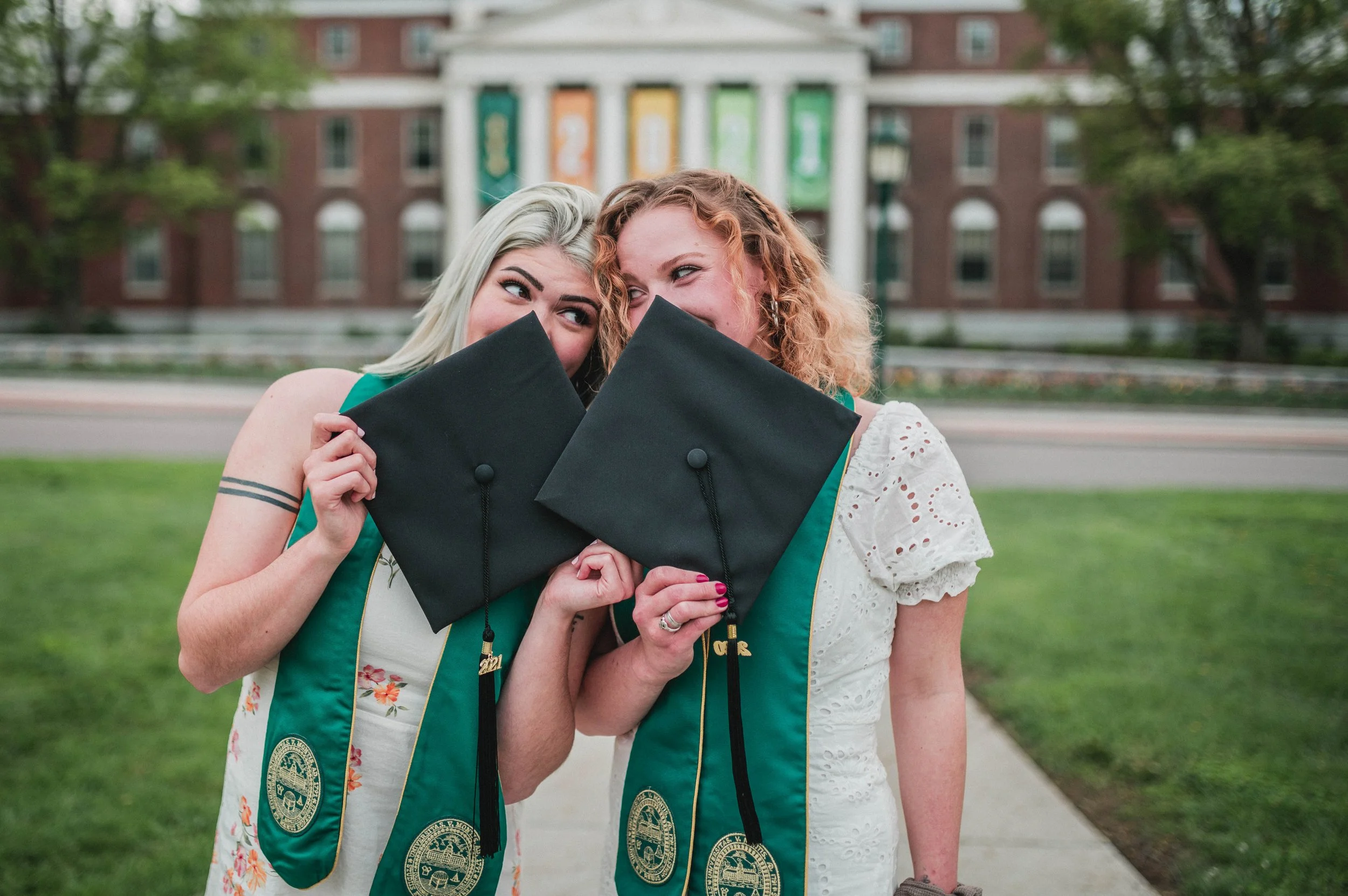 Two graduates holding caps in front of a building with columns and banners, wearing green stoles and dresses.