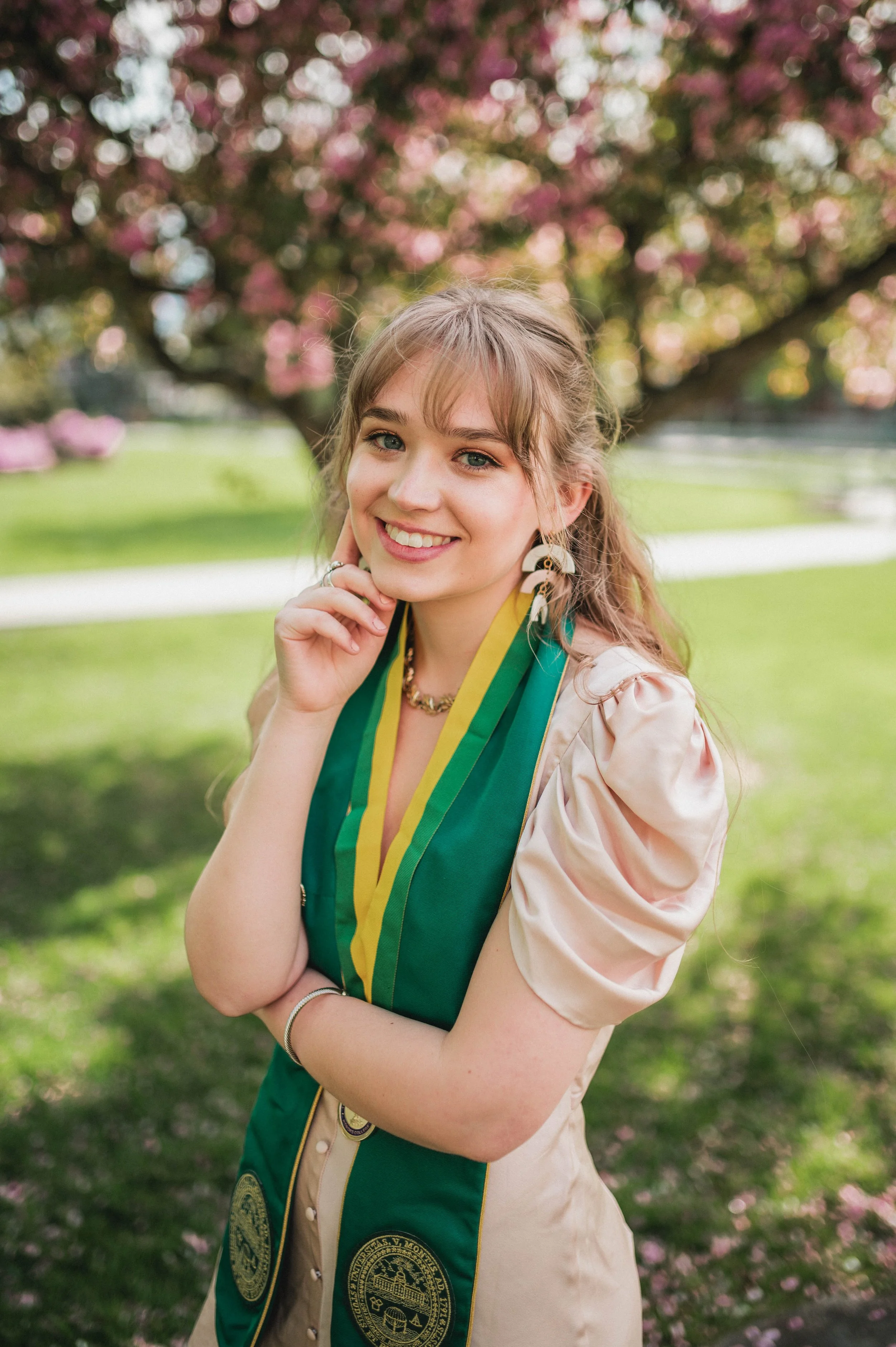 Young woman wearing a green graduation stole and a light-colored dress, smiling in a park with blooming trees in the background.