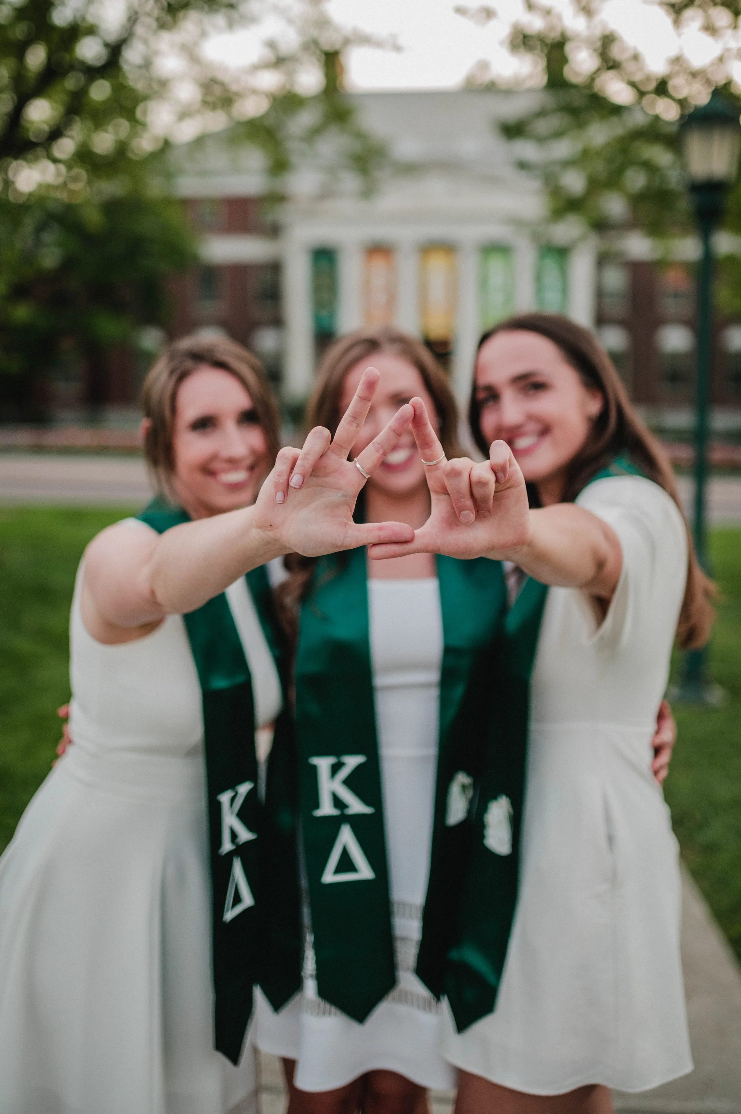 Three women in white dresses wearing green Kappa Delta stoles, posing with a hand gesture, smiling outdoors on a college campus.