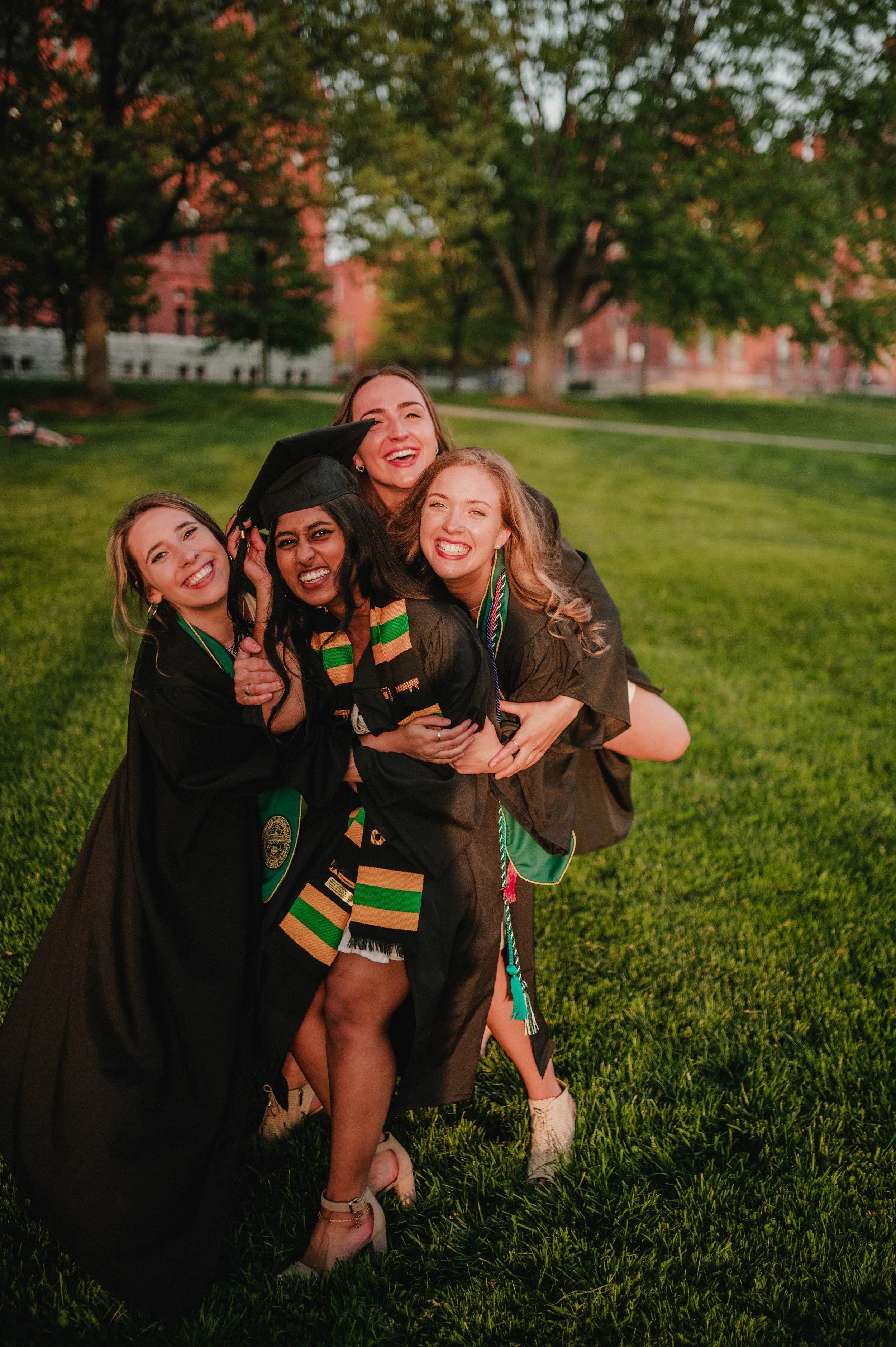 Four graduates in caps and gowns celebrate and hug on a grassy lawn.