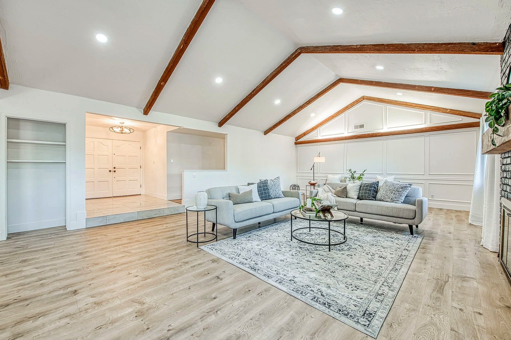 Bright living room with vaulted ceiling, wooden beams, white walls, two gray sofas, a patterned rug, round black coffee tables, and modern ceiling lights.