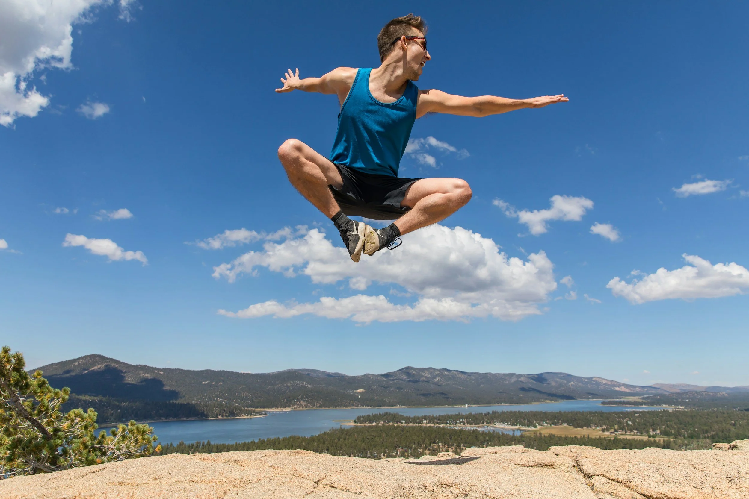 A man wearing sunglasses, a blue tank top, and black shorts is jumping in the air with his arms and legs outstretched against a backdrop of mountains, a river, and a partly cloudy sky.