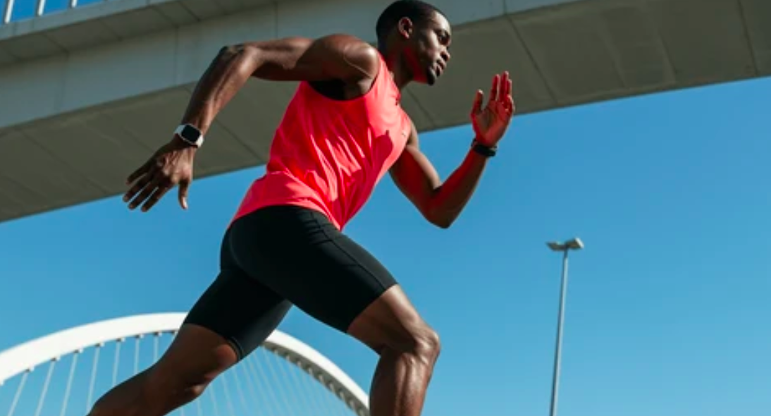 A male runner in a red shirt and black shorts is sprinting outdoors under a bridge on a sunny day, showing energy and stamina after vitamin b12 injection.