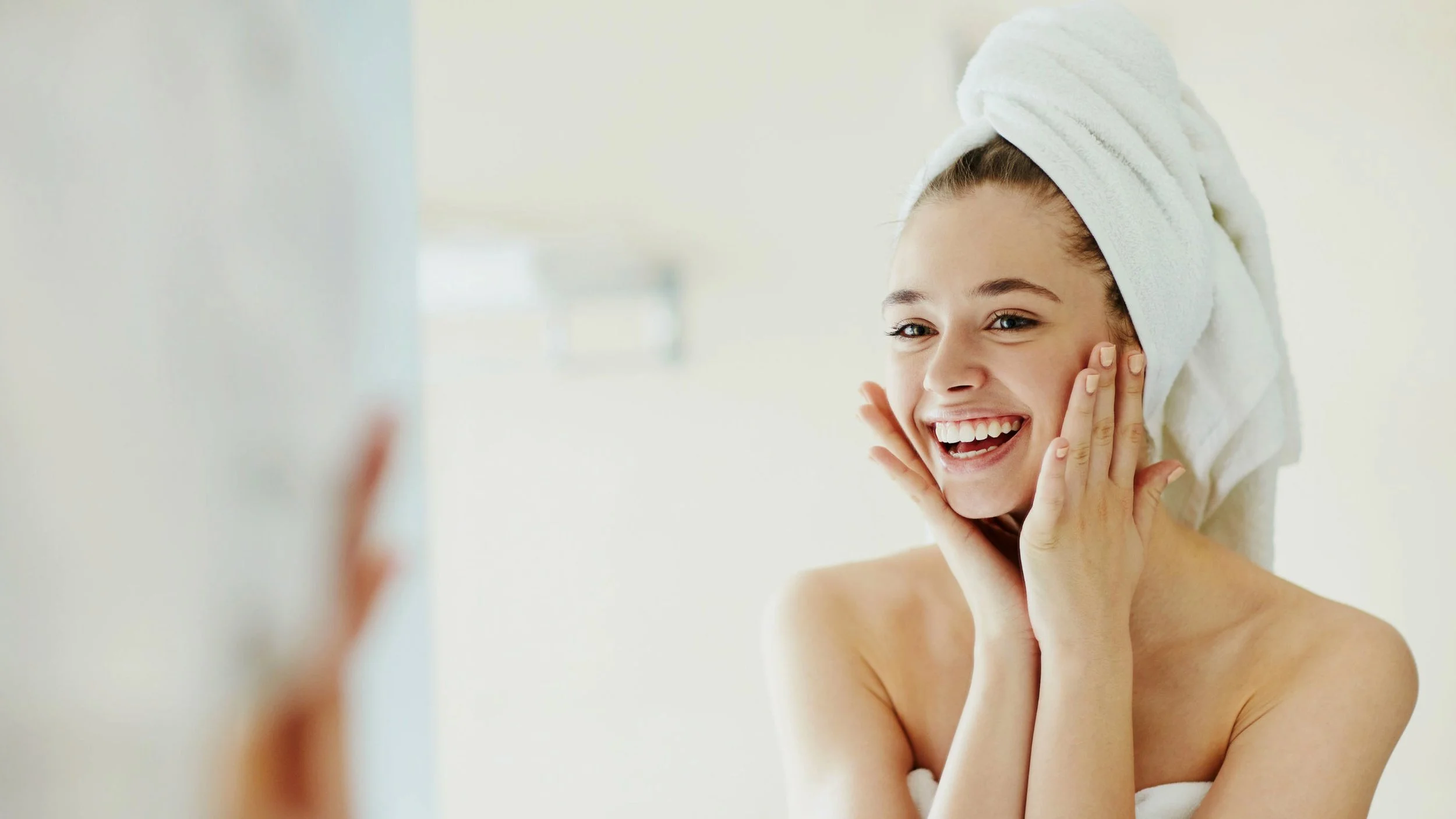 Young woman with a towel wrapped around her head, smiling and touching her face, in a bright bathroom.