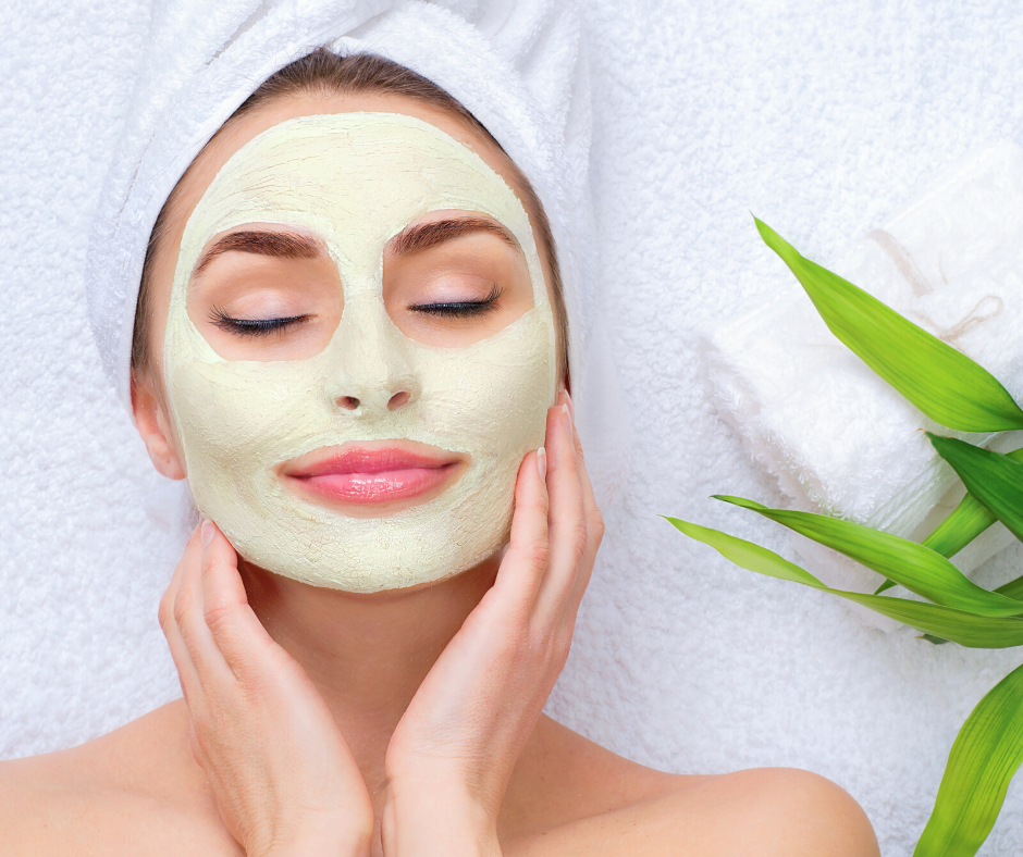A woman lying on a towel with a facial mask applied, eyes closed, smiling gently, with green leaves nearby.