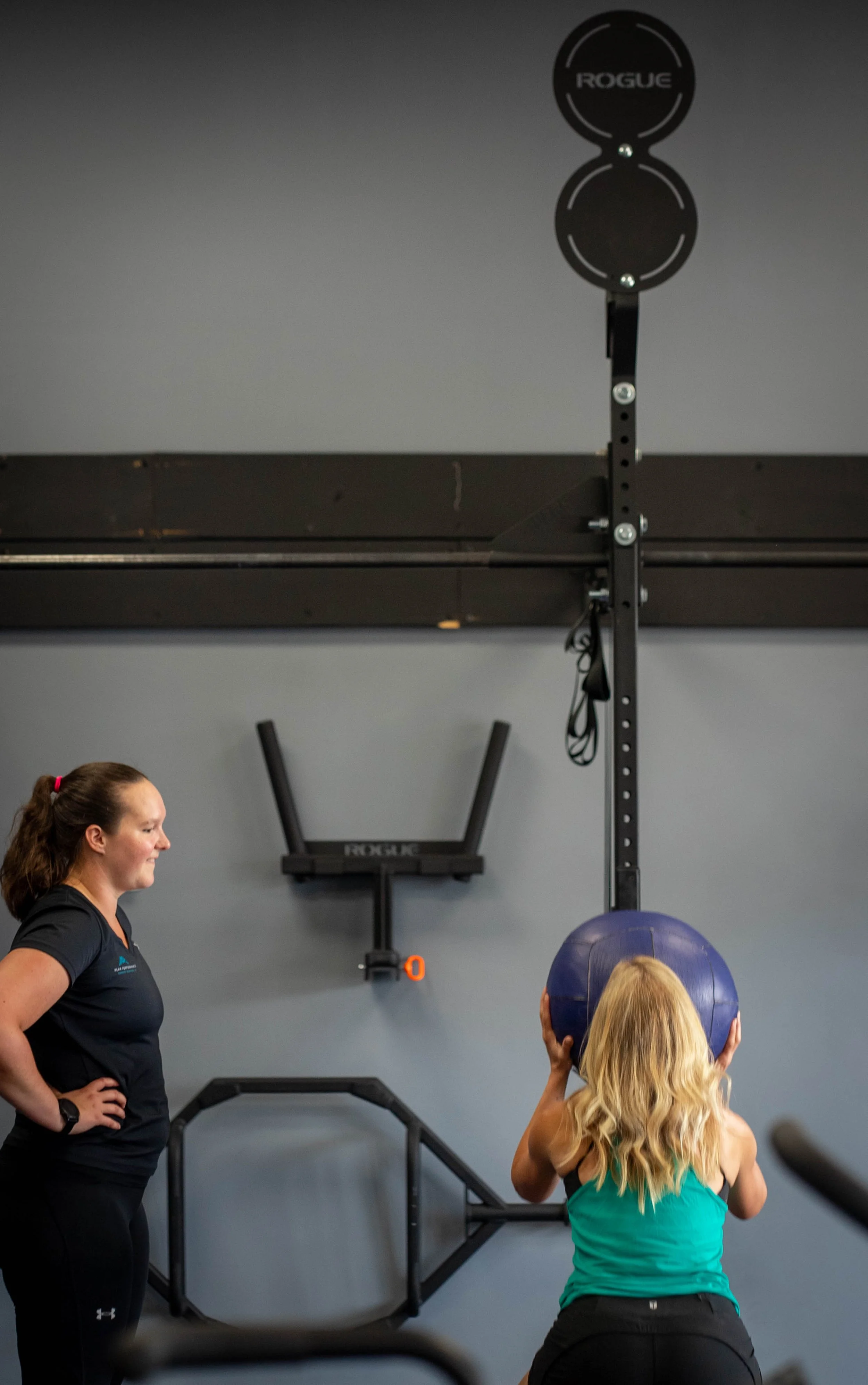 A woman in a blue tank top holding a medicine ball in a gym, with a trainer standing beside her, and workout equipment like a pull-up bar and wall-mounted accessories visible in the background.