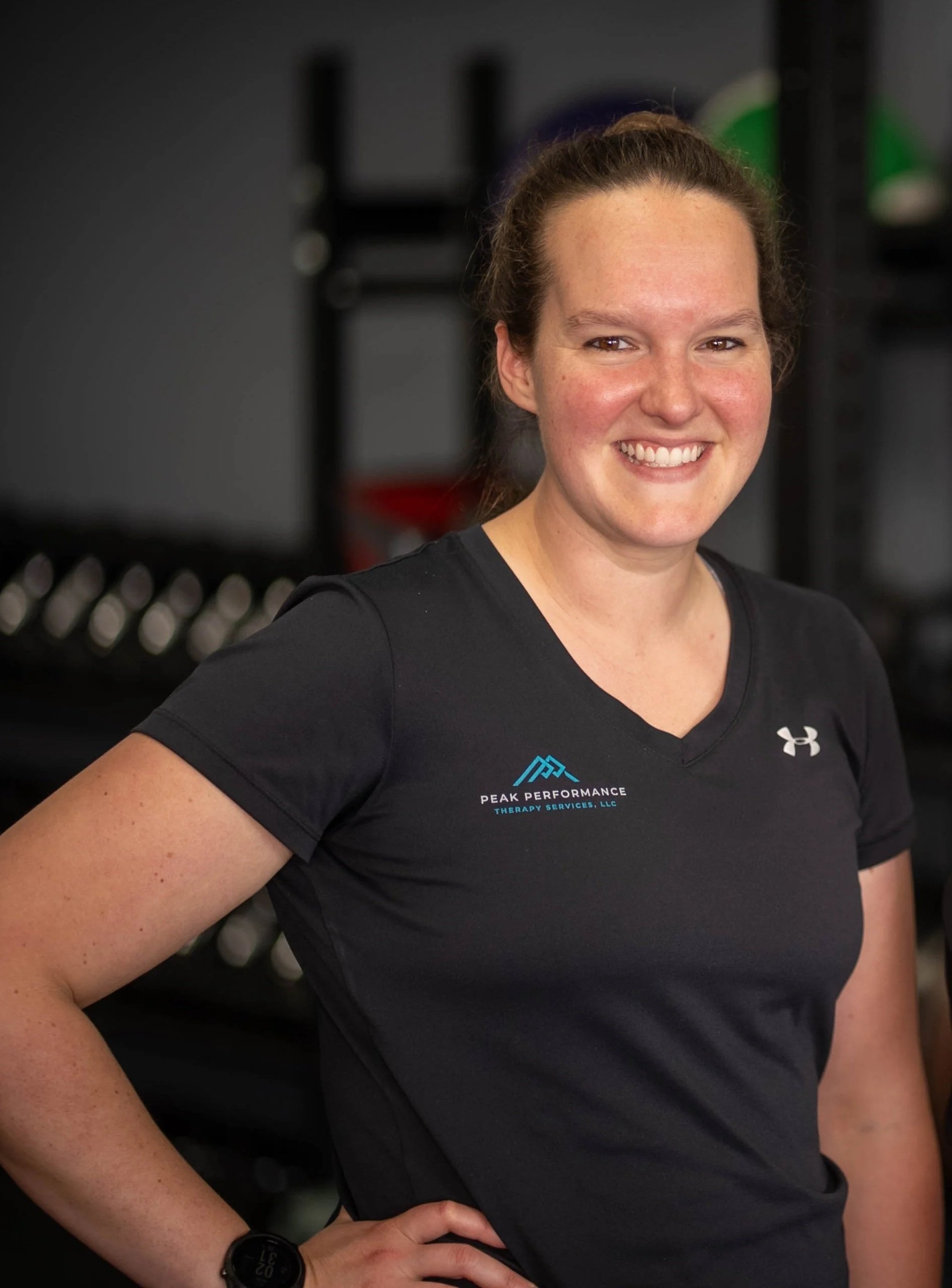 A smiling woman with brown hair in a ponytail, wearing a black athletic shirt with Under Armour logo, standing with crossed arms in a gym with dark background and exercise equipment.