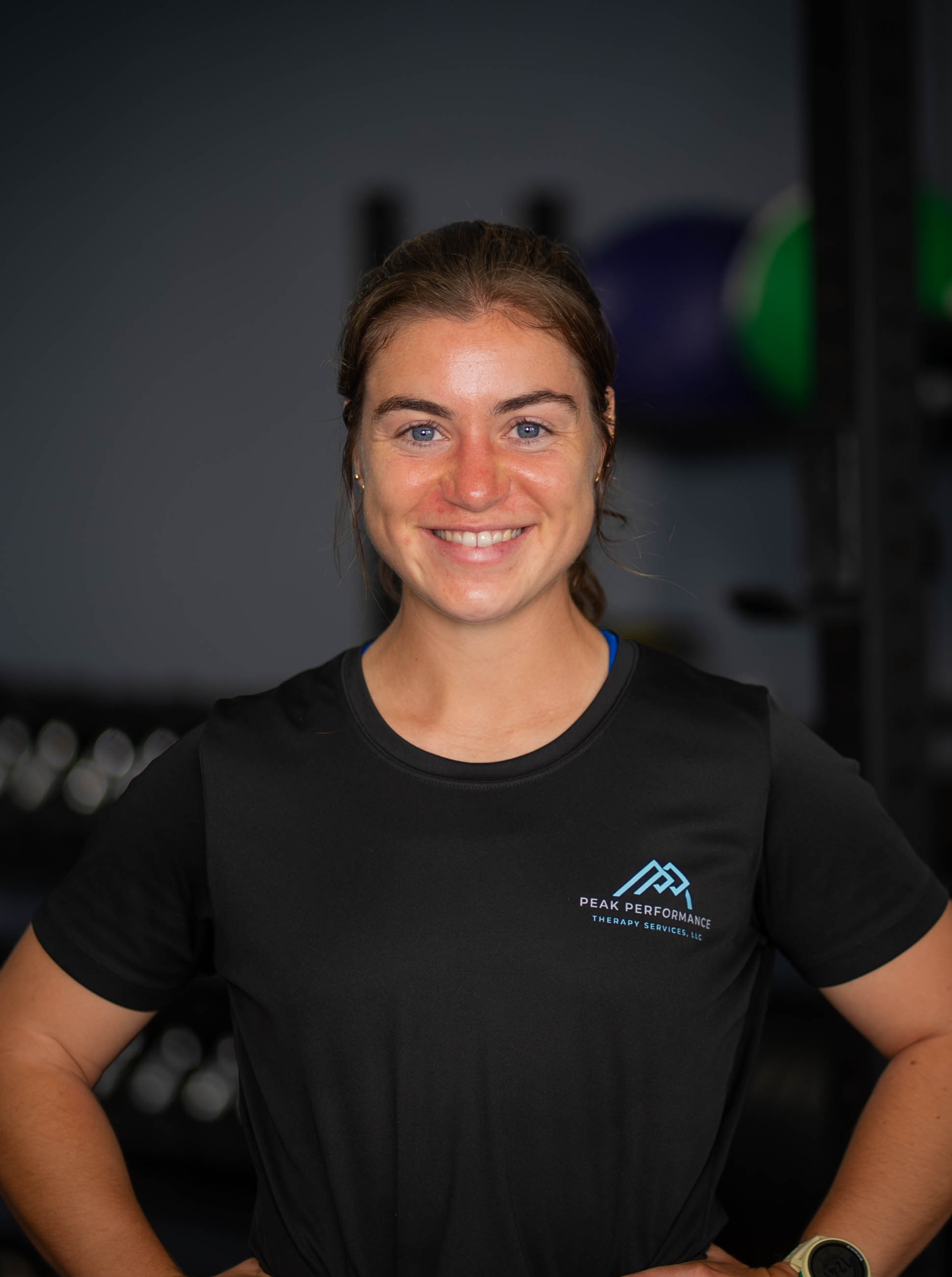 Smiling woman wearing a black Peak Performance Therapy Services LLC T-shirt, standing in a gym with exercise equipment in the background.