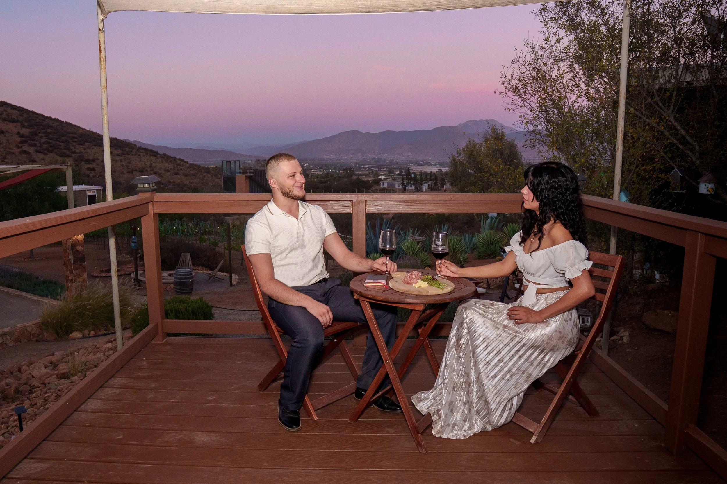 A man and woman sharing a romantic dinner on a wooden deck with a mountain view at sunset. They are holding glasses of red wine and smiling at each other. The table has a charcuterie board with meats, cheese, and herbs.