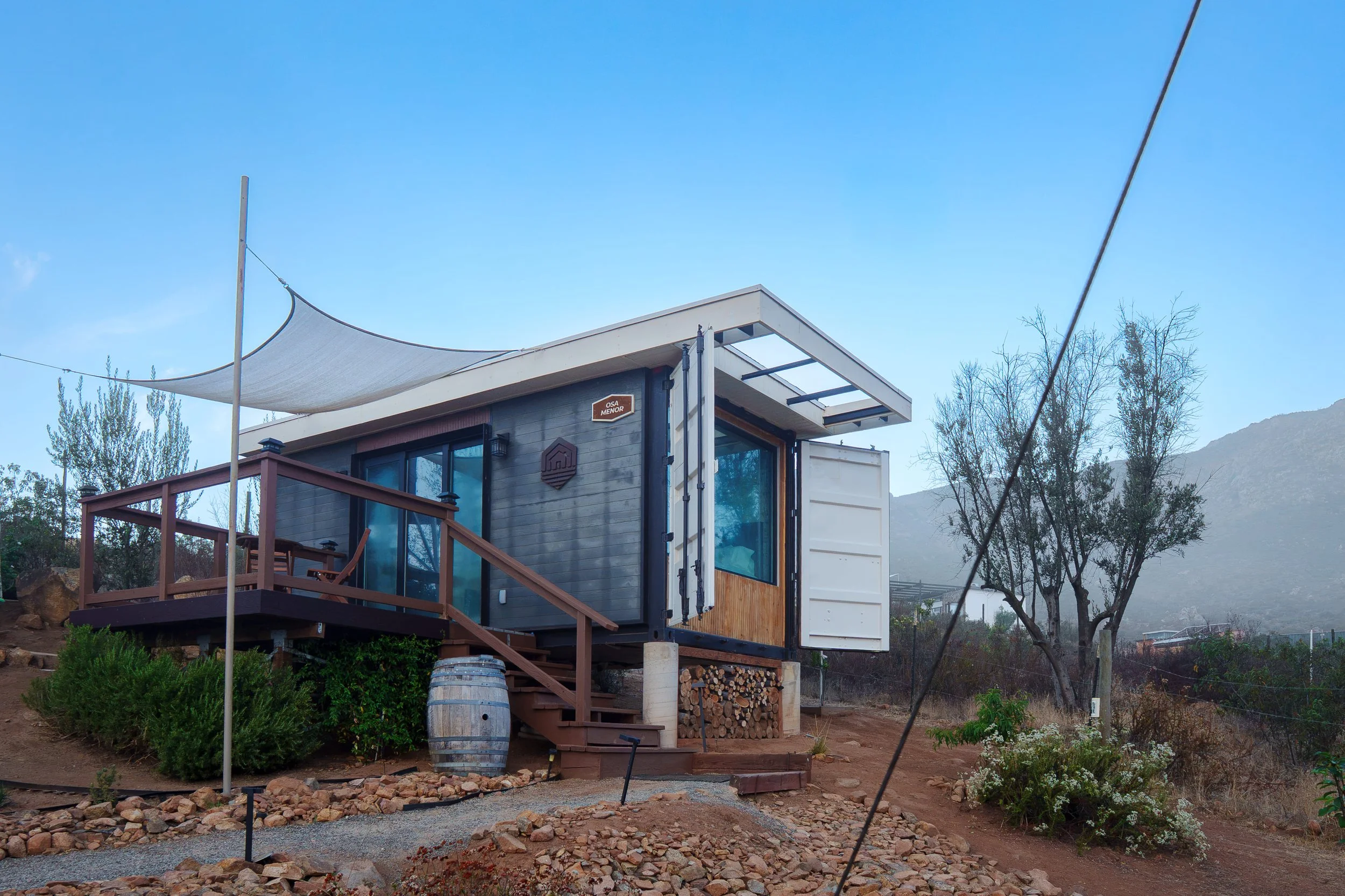 Modern tiny house built on a trailer with a small patio, stairs, and wood storage underneath, surrounded by plants and trees, with a mountainous background and blue sky.