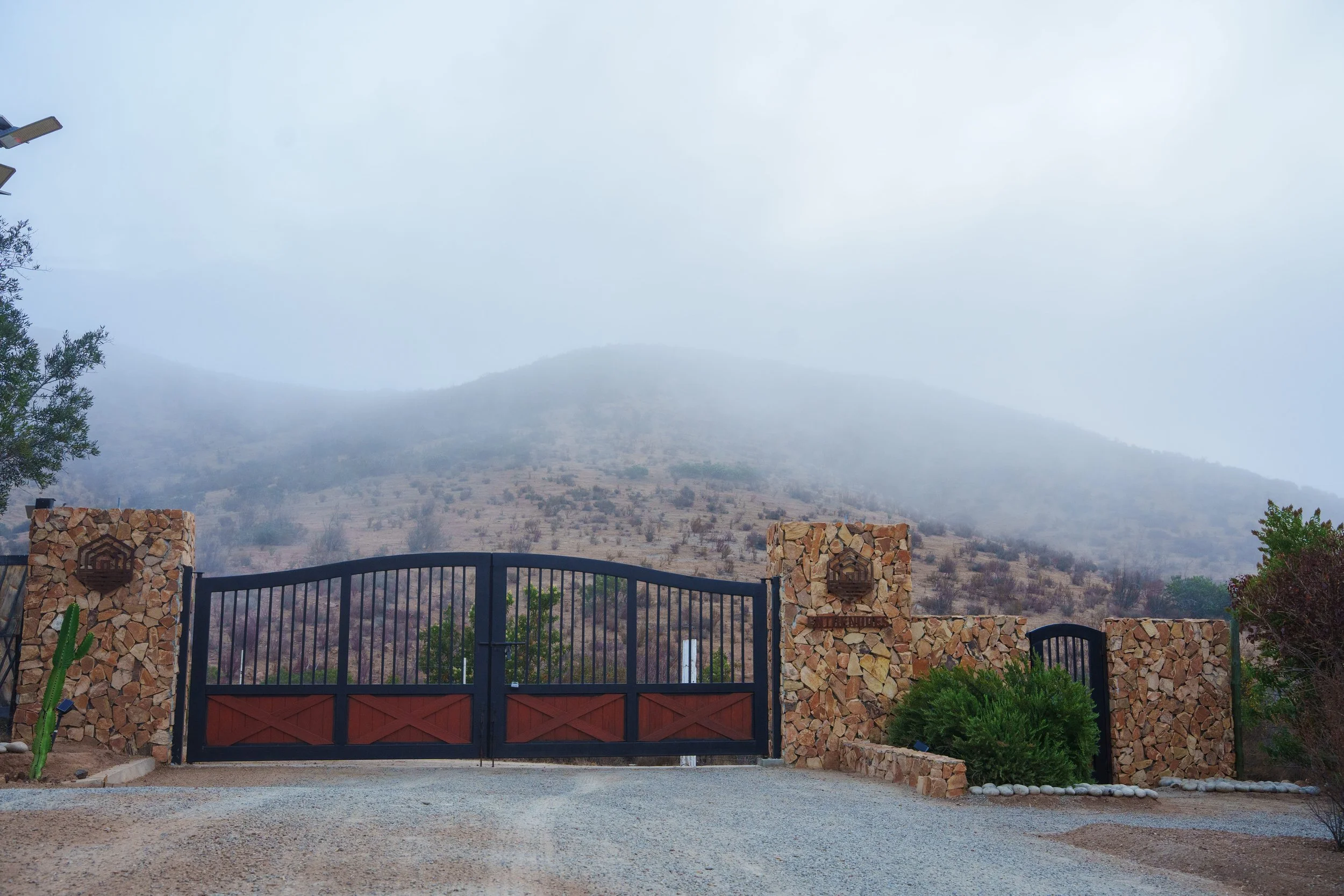 View of a landscape with mountains partially obscured by fog, a stone and metal gate in the foreground, and desert vegetation surrounding the area.