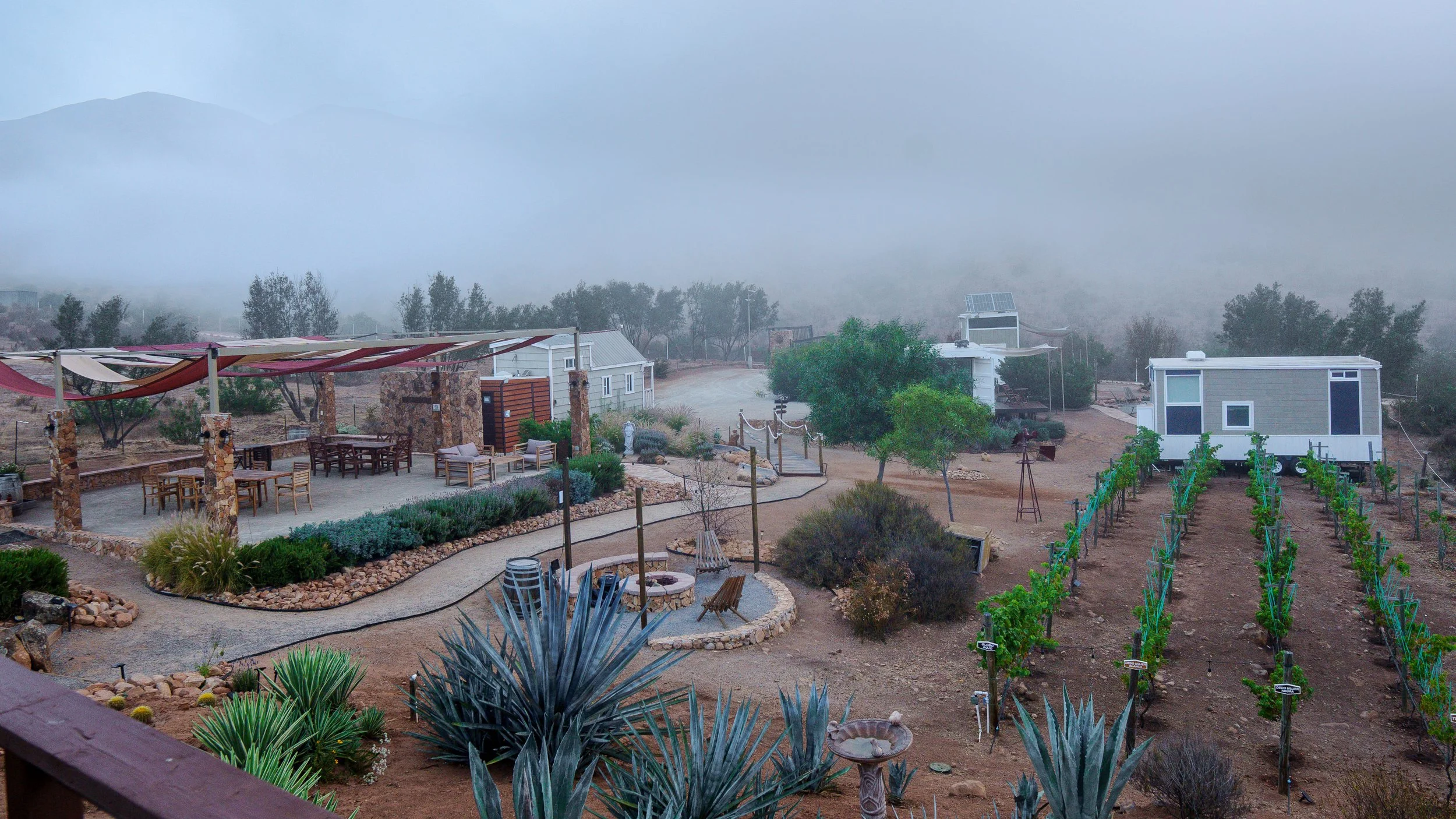 A desert landscape with small modern mobile homes, outdoor furniture, trees, plants, and a foggy mountain in the background.