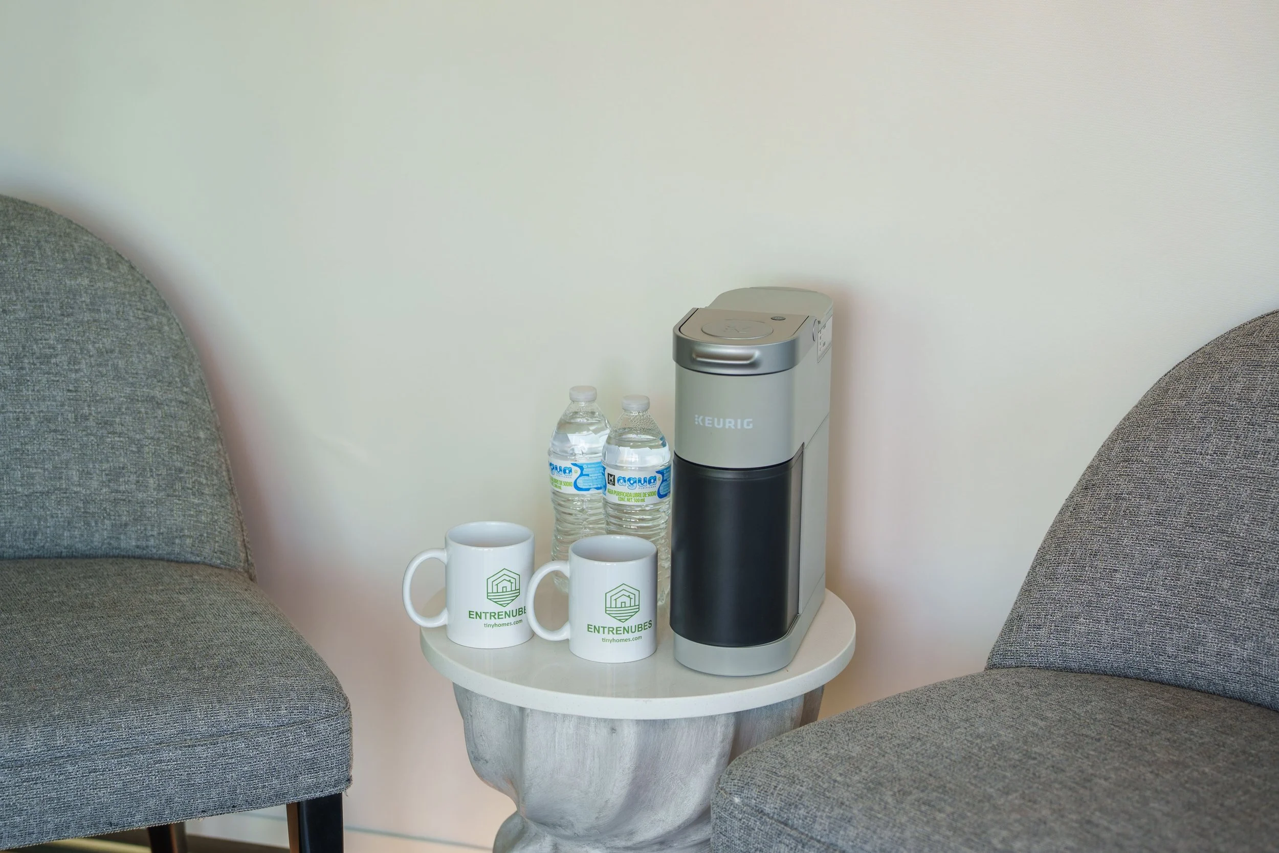 Two gray upholstered armchairs facing each other with a small white round table between them. On the table are two white mugs with green logos, two bottles of sparkling water, and a gray water dispenser. The background features a plain white wall.